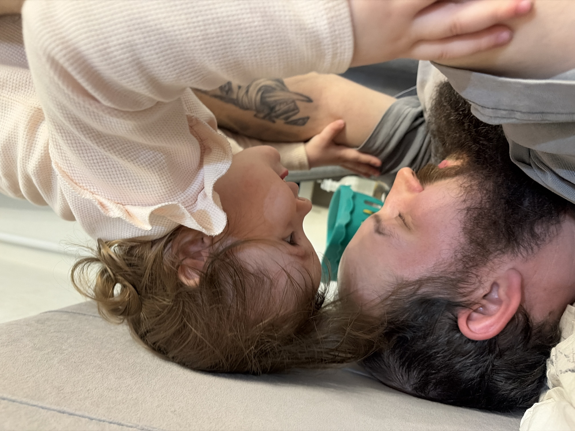 A close-up, angled view shows a man with a dark beard and a child, lying head-to-head on a light-colored surface, possibly a couch or bed. The man's face is close to the camera, partially obscured by his beard. The child, wearing a light-colored, ruffled top, is angled upwards with their head tilted. An adult hand, likely belonging to another person, rests on the man's arm, which is visible in the background and has a tattoo. The background features muted tones and indistinct objects, including a turquoise object. The location is Chaikovsky, Russia.