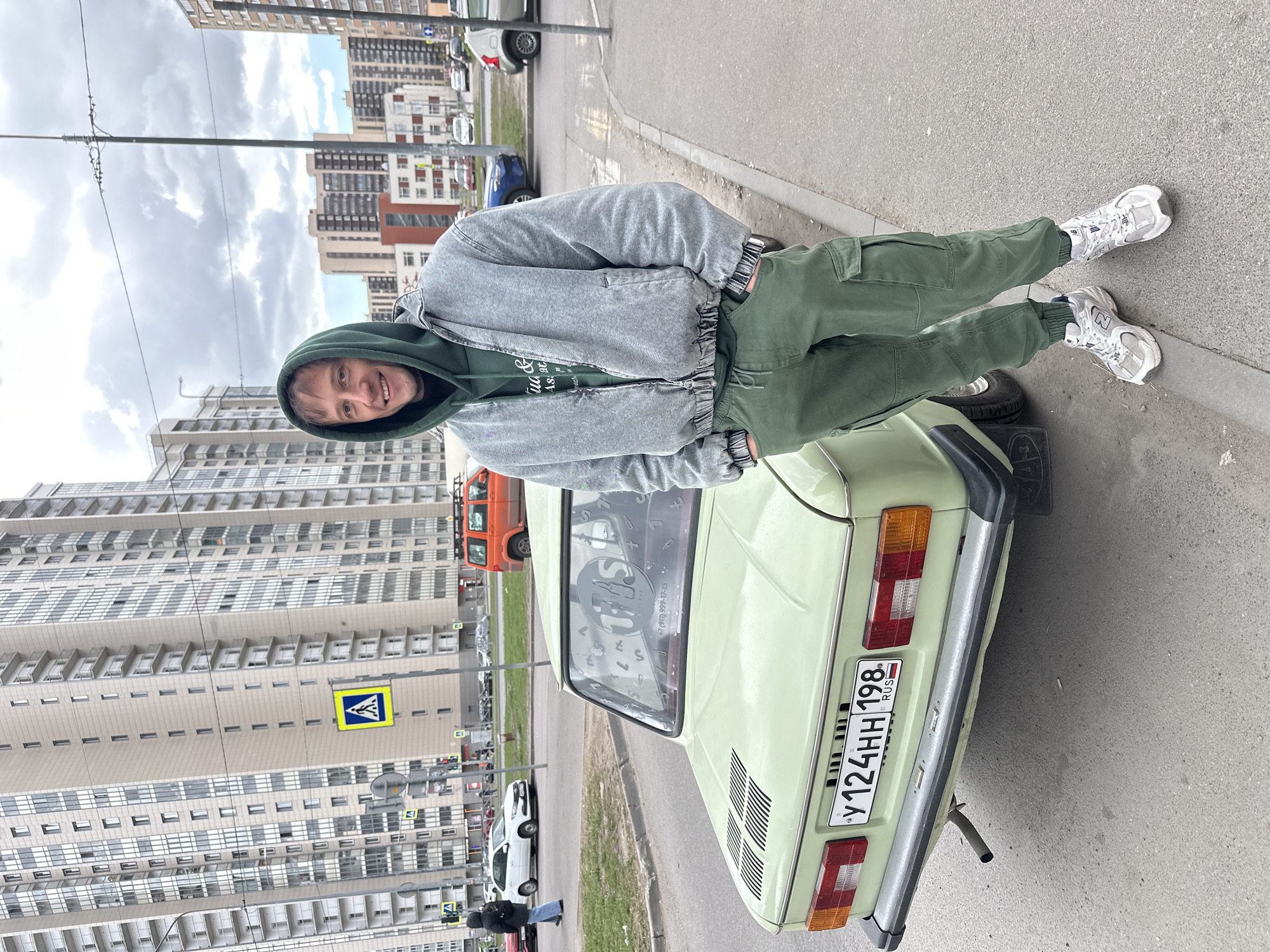 A young man is posing next to a light green vintage-style car on an overcast day in Murino, Russia. He is smiling and looking towards the camera. The man is wearing a dark green hooded sweatshirt, partially revealing text like "qua & As at," a light gray distressed jacket, olive green cargo pants, and white New Balance 530 sneakers.
The car, an older model, has a Russian license plate "Y124HH 198 RUS" on its rear. A sticker on the back window displays the word "SHAS," a phone number "+7 (911) 999-17-15," and various other letters and symbols. The vehicle is parked alongside a road with a paved sidewalk.
In the background, several tall, modern residential buildings dominate the skyline under a cloudy sky. Street furniture, including power lines, poles, and a pedestrian crossing sign, are visible. Other cars, including a white sedan and a distant orange truck, are parked along the street. The scene depicts a casual moment in an urban residential area.