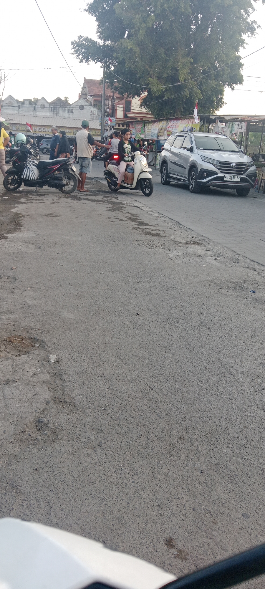 The image depicts a street scene in Maguwoharjo, Indonesia, likely a commercial area given the buildings and businesses visible. A silver SUV with license plate "AB 1289 EP" is parked on the right side of the road, facing forward. In the middle ground, a woman is riding a white scooter, and behind her, another scooter is parked. Several people are standing or walking around, some near parked motorcycles. In the background, there are buildings with awnings and signs, including one that reads "INDOMIE." A large tree with dense foliage is prominent in the upper right portion of the frame, casting shadows on the scene. The ground is paved asphalt, showing signs of wear and some debris. The sky is overcast.