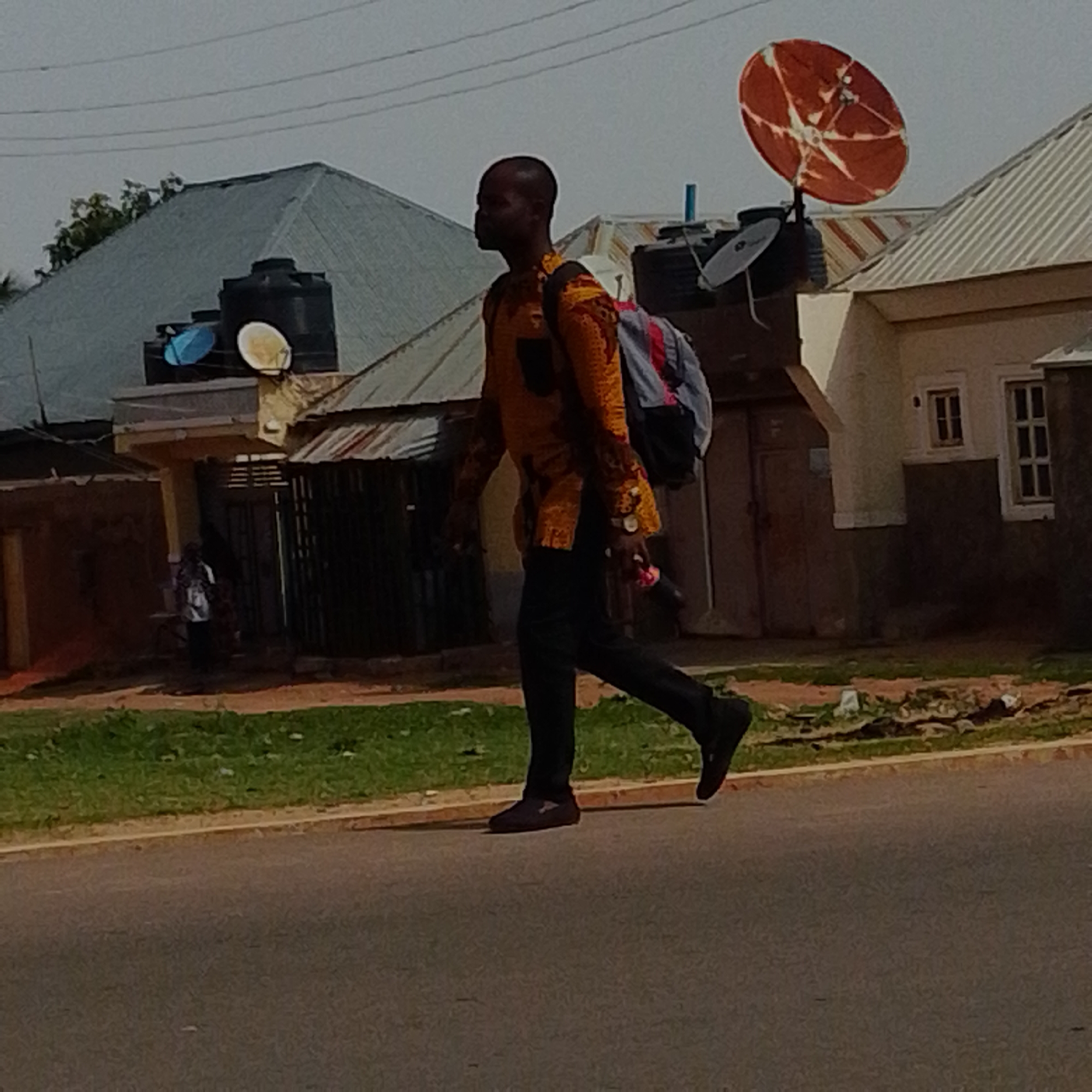 A man walks across the street in Gombe, Nigeria, wearing a brightly patterned yellow and orange shirt, black pants, and black shoes. He carries a backpack and a red object in his right hand. In the background, there are buildings with corrugated metal roofs and satellite dishes. Some of the buildings appear to have water tanks on top. A small child can be seen standing near a doorway in the background. The ground in front of the buildings is covered in grass and dirt. The scene suggests a residential or commercial area within the city.