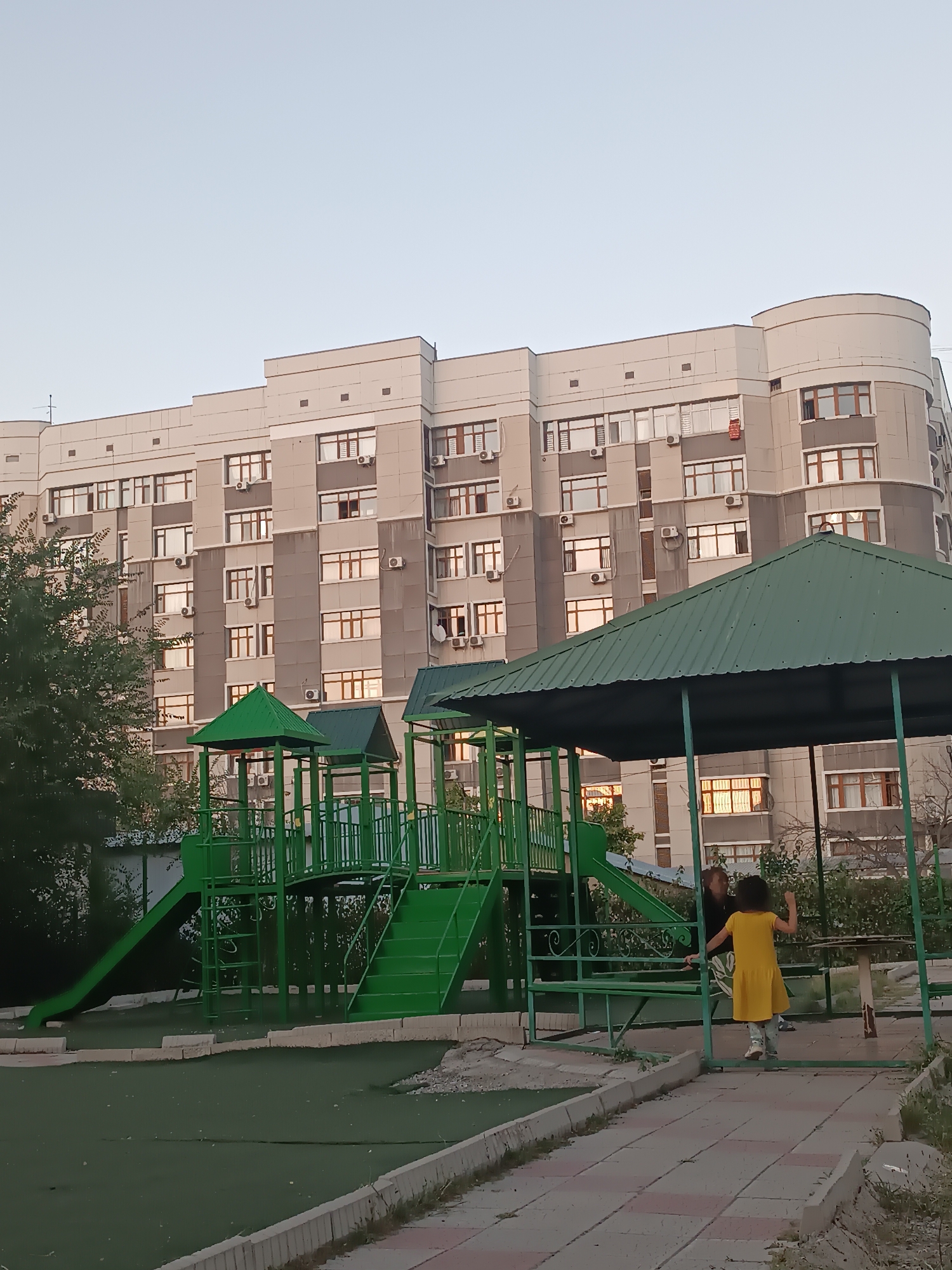 A girl in a yellow dress stands on a paved walkway near a green playground structure. The playground has a slide and stairs. Behind the playground is a large, modern apartment building with many windows. The sky is a pale blue, and the lighting suggests it is either early morning or late evening. The setting appears to be an urban residential area.