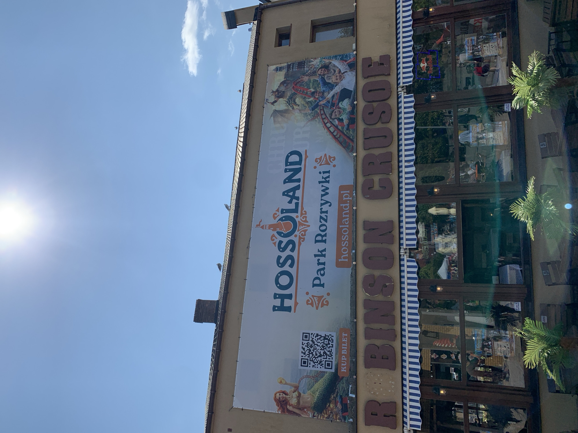 The image shows the facade of a building with a large banner advertising "HOSSOLAND Park Rozrywki". Below the banner, the name "ROBINSON CRUSOE" is prominently displayed in large, brown, three-dimensional lettering. The building has a brown exterior with large windows framed by wooden beams. A blue and white striped awning stretches across the top of the windows. In the windows, reflections of trees and people are visible, as well as posters and what appears to be a Red Bull neon sign. In front of the building, there are tables and chairs, and potted palm plants. The sky above is clear blue with a few scattered clouds, and the sun is bright, causing some lens flare. The building is located in Rewal, Poland, as indicated by the banner.