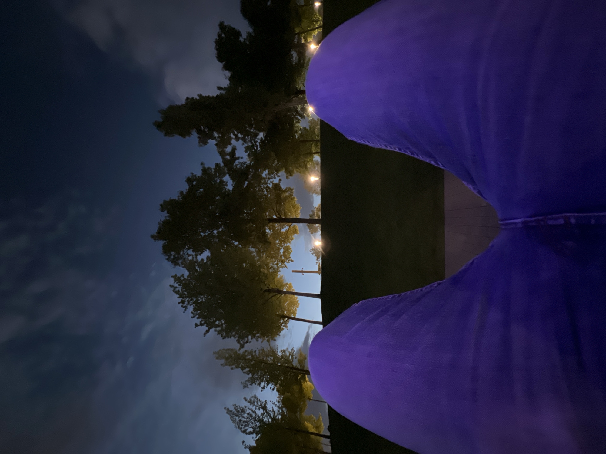 The image, taken at night in an outdoor setting, features a pair of blue legs positioned in the foreground, likely belonging to a seated person. Beyond the legs, the scene depicts trees illuminated by strung lights. The sky is a deep blue with some visible clouds. The location, Krasnodar, Russia, cannot be confirmed based on visual evidence alone.