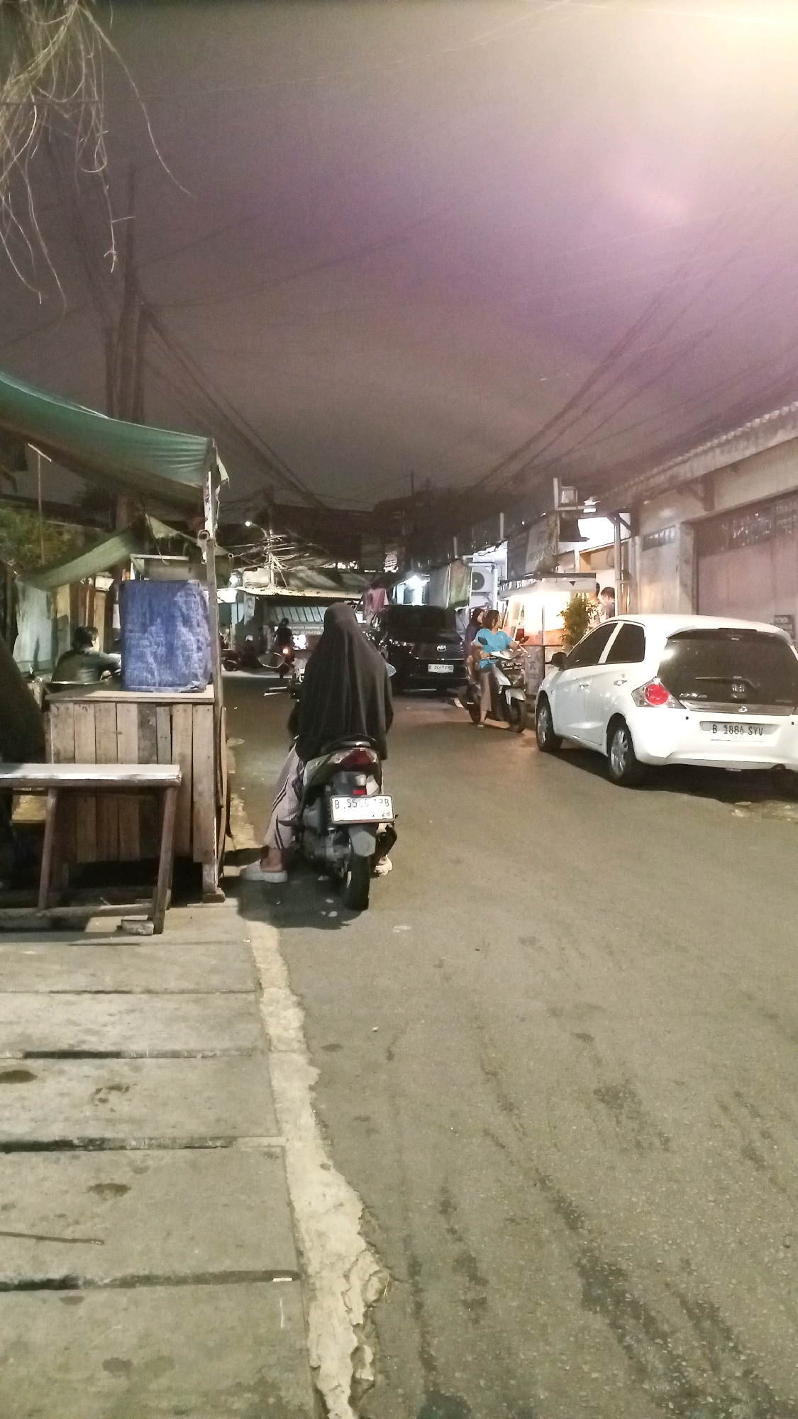 This is a nighttime, medium-shot photograph taken on a street in South Jakarta, Indonesia. The scene depicts a narrow urban road with a sidewalk to the left and buildings lining the right. Overhead, a dense network of electrical wires is visible against a hazy, dimly lit sky with a purplish hue.
In the foreground, a person wearing a black hijab and striped pants is seated on a dark-colored scooter. The scooter's license plate reads "B 5506 TPH." To the left of the scooter, a wooden stall with a blue tarp is partially visible, with another person seated behind it. The sidewalk is paved with concrete slabs, some of which appear worn and cracked.
Further down the street, a white car is parked on the right side; its license plate is "B 1686 SYU." To the left of the white car, a black car is also parked. Another scooter is moving away from the camera on the right side of the road, ridden by a person wearing a blue top. The street itself is asphalt, showing signs of wear and scattered debris. Buildings with awnings and open fronts are visible on the right side of the street, illuminated by artificial lights, suggesting commercial activity. The overall atmosphere is that of a bustling but somewhat unkempt urban environment at night.