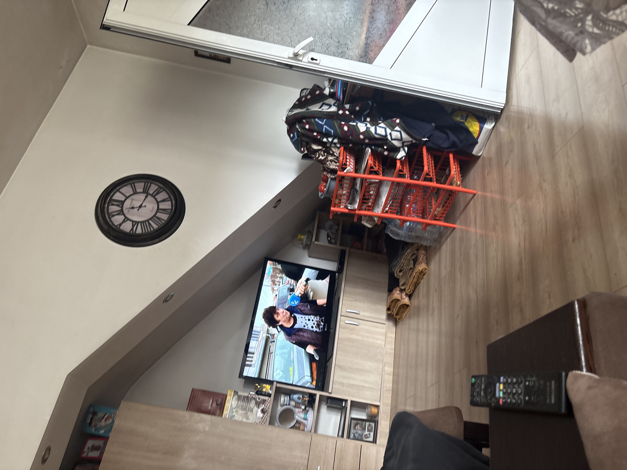 The image presents an interior view of a room, likely a living space, in Staro Selo, Bulgaria, recognizable by the furniture and items within. A prominent feature is a wall-mounted television displaying a news broadcast with a reporter present. Below the TV, there is a shelf unit containing various decorative objects. On the wall, a large clock is visible. The furniture includes a sofa or seating arrangement, part of which is visible in the foreground along with a remote control. The environment seems like a private residence.