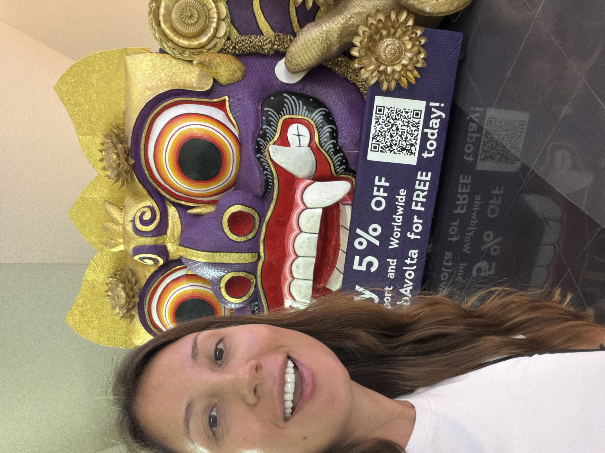 A woman stands in front of a large, purple and gold decorative mask, likely a Barong mask, in Kuta, Indonesia. The mask features exaggerated eyes with concentric rings of red, orange, and yellow, a gaping mouth with large white teeth, and ornate gold headdress elements. Below the mask, a purple banner displays text and a QR code. The woman is smiling and looking at the camera, with her shoulder and chest visible, wearing a white t-shirt. The floor is dark and reflective, showing a distorted reflection of the banner and QR code. The lighting suggests an indoor environment, possibly a shopping mall or cultural center, given the ornate display.