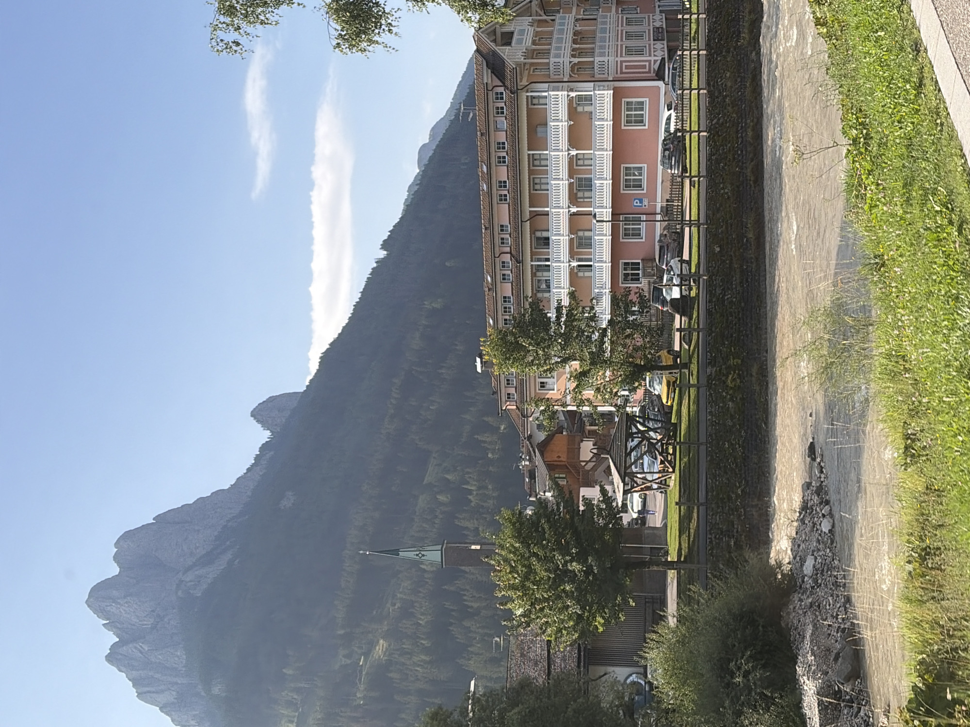 The image depicts a scenic view of Pozza di Fassa, Italy, with a prominent mountain range in the background. A tall, slender church steeple with a green spire rises above the rooftops of a charming town. A multi-story pink building with white balconies and numerous windows dominates the middle ground, suggesting a hotel or residential complex. In the foreground, a fast-flowing river, the Avisio, meanders through the landscape, its waters a muddy brown color. The riverbanks are lined with green grass and some rocky debris. Several cars are parked along the street in front of the pink building, and a wooden gazebo-like structure stands near the river. Trees are scattered throughout the scene, adding to the natural beauty of the location. The sky is a clear blue, with a few wispy clouds.