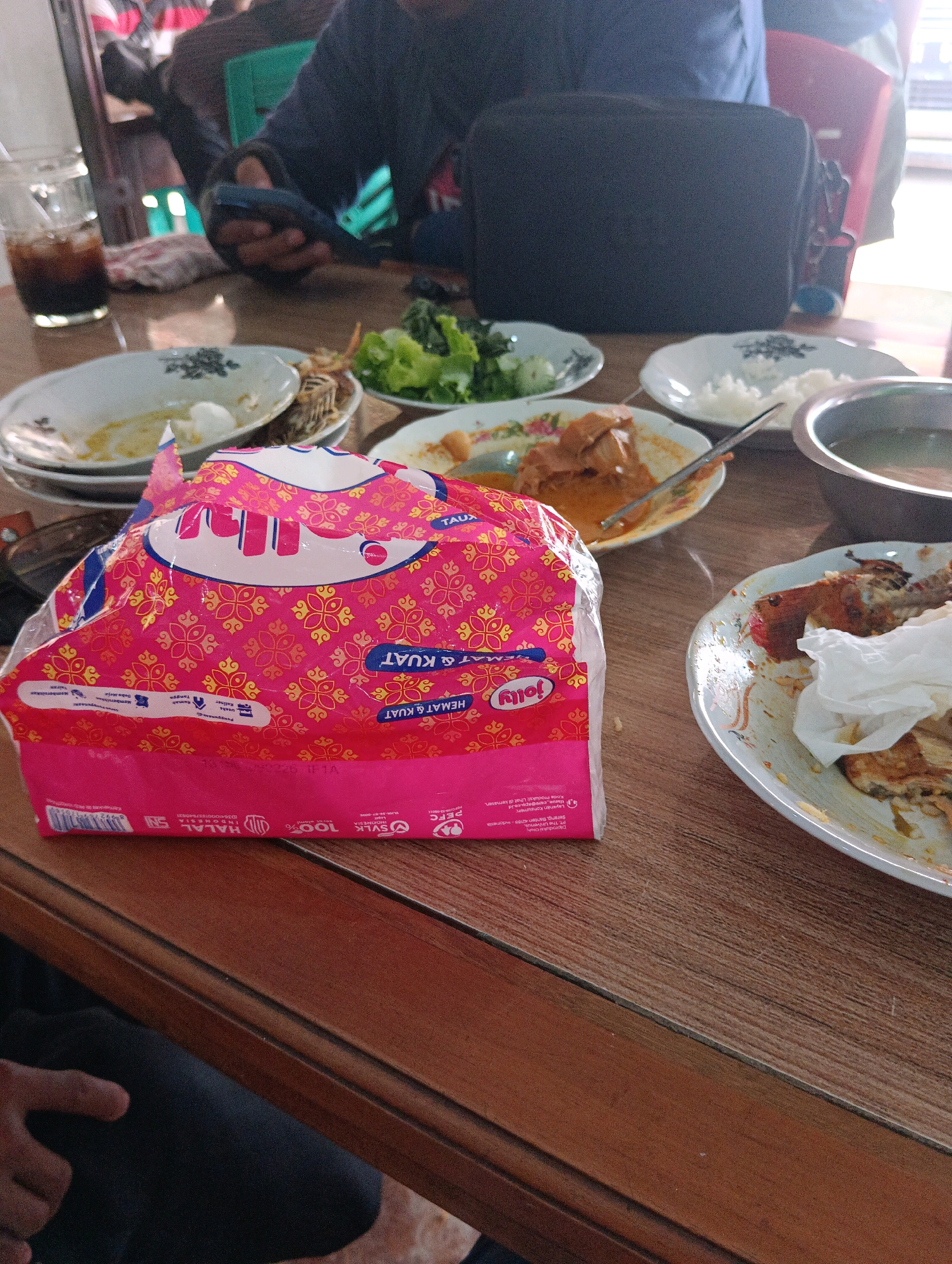 A medium shot captures a wooden table laden with food and drinks, set for a meal. A pink and red patterned package of "Jolly" brand tissues is prominently positioned in the foreground. Several plates contain remnants of food, including what appears to be rice, chicken, and vegetables. A glass filled with a dark beverage is on the left, and a bowl of soup is on the right. In the background, out of focus, are people seated, suggesting a casual dining setting. The scene appears to be an indoor establishment, likely a restaurant or a home, in Belitang, Indonesia.