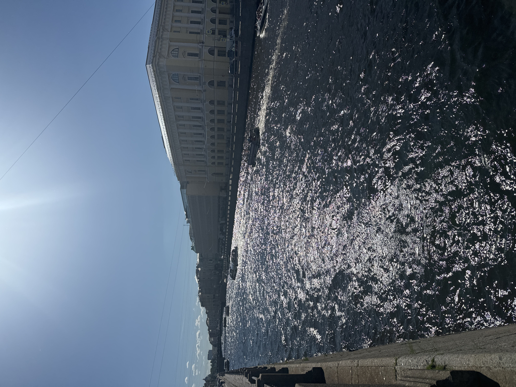 This media file captures a view of a wide river in Saint Petersburg, Russia, on a bright, sunny day. The river is dark and choppy, with sunlight glinting off the ripples, creating a shimmering effect. Along the riverbank, a long, light-colored building with many windows stretches into the distance, its classical architecture suggesting a historic district.
Several small boats are visible on the water, with one in the middle ground appearing to be a tour boat with passengers. The sky is a clear, bright blue, with a few wispy clouds in the distance on the right side of the frame. The sun is high, indicating it's likely midday. The scene evokes a feeling of a pleasant, warm day in a European city. A concrete embankment with a stone railing runs along the foreground, providing a vantage point for observing the river and the cityscape. Some electrical wires are also visible crossing the sky.
