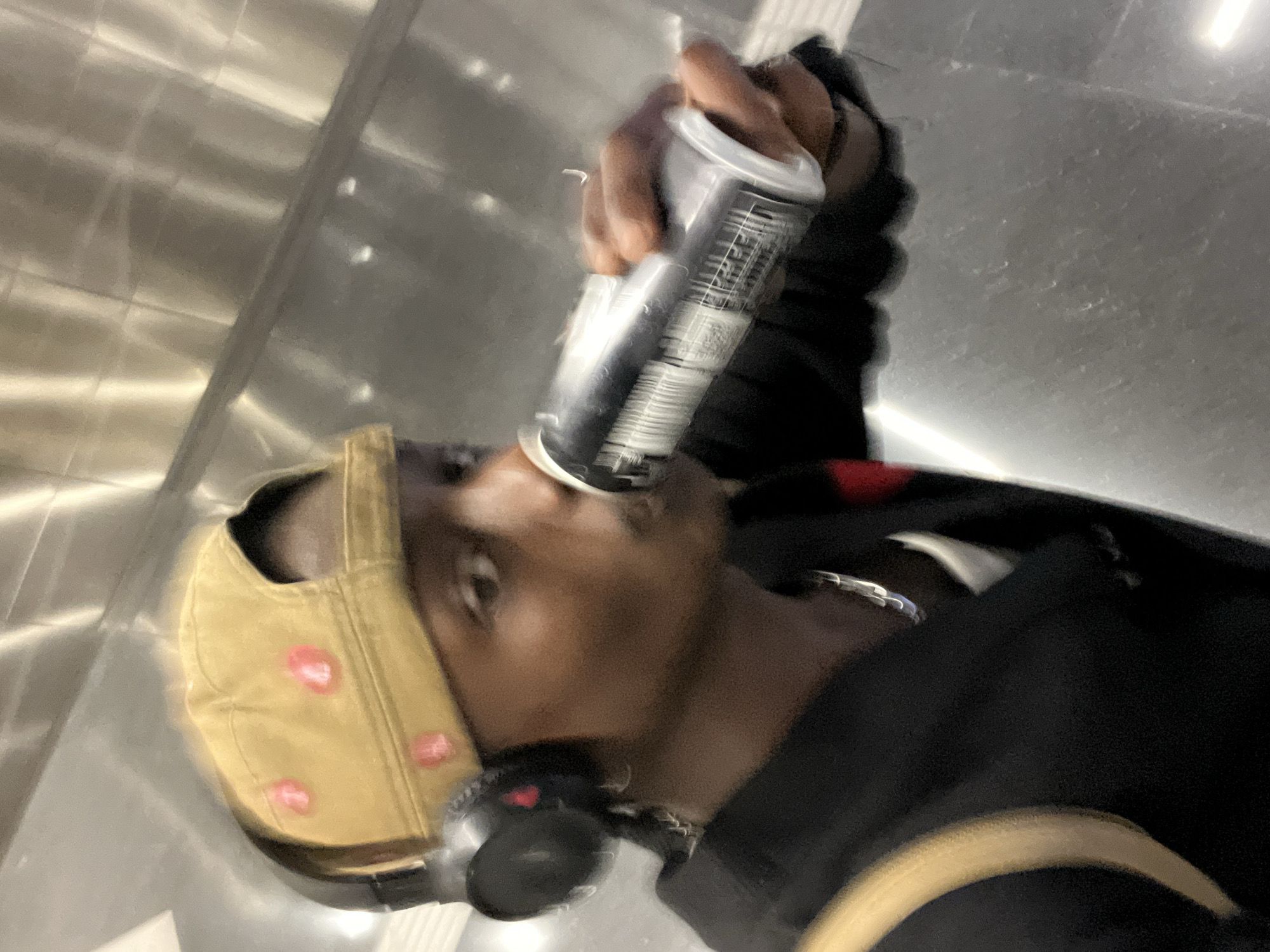 An indoor, motion-blurred portrait shot captures a person drinking from a silver-colored can. The image is rotated clockwise, presenting the subject and background at an angle. The person, who has dark skin, is seen in profile, holding the can directly to their mouth. They are wearing a light brown or tan baseball cap adorned with several pink circular details, black headphones around their neck, and a silver-colored chain or necklace. A dark-colored jacket or hoodie is also visible.
The can itself is silver with heavily blurred labels, preventing any text, branding, or specific product identification. The entire scene is affected by significant motion blur, making precise details indistinct and suggesting movement at the time the photo was taken. The background consists of shiny, reflective panels, possibly metallic or brushed steel, indicative of a modern interior space, such as an elevator or contemporary building hallway. The lighting appears artificial, reflecting off the glossy surfaces. Due to the blur, facial features and specific emotions are not discernible. No legible text is visible on any object.