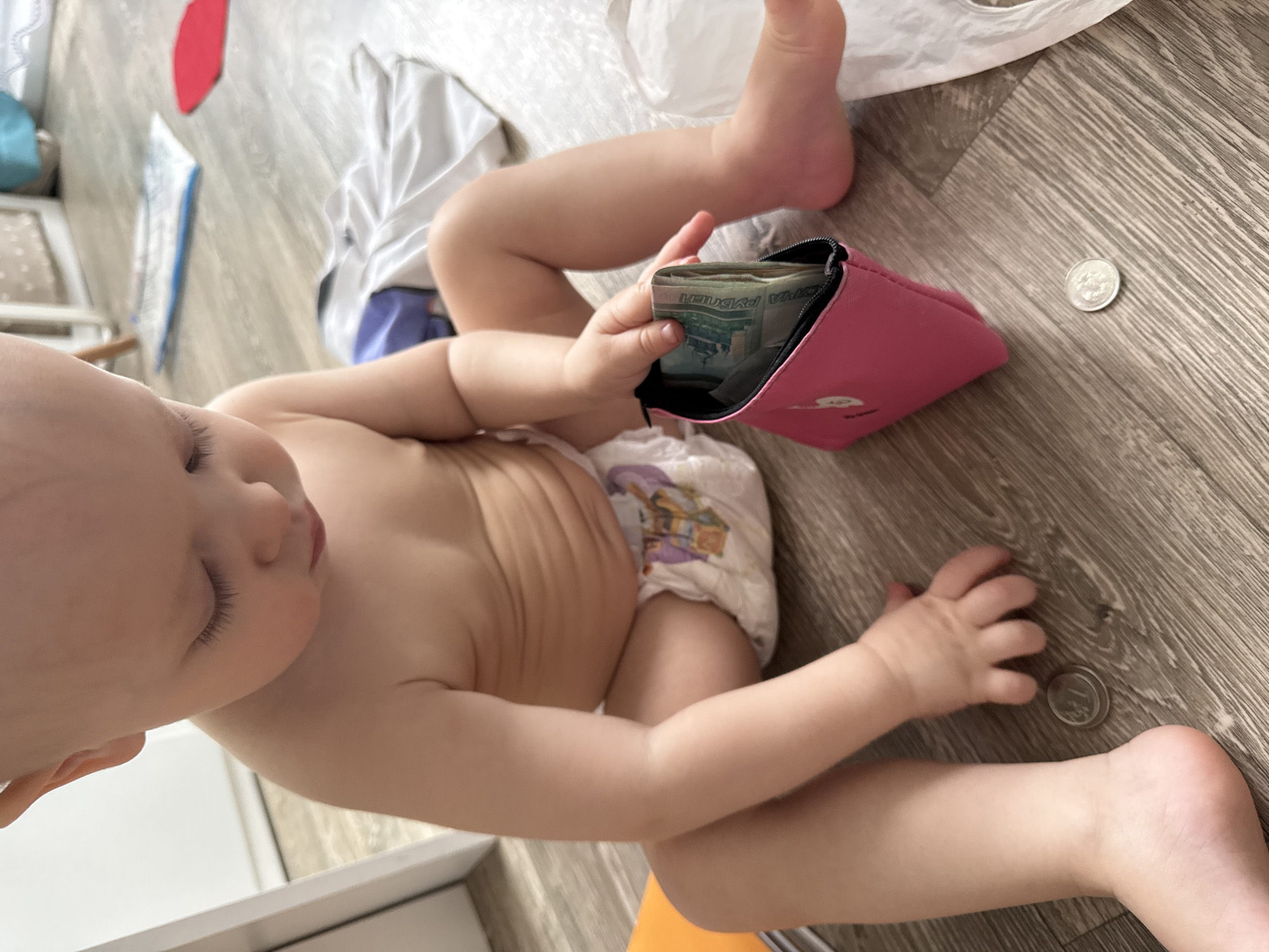 A baby, wearing a diaper, is sitting on a wooden floor, holding a partially open pink wallet filled with paper money. Next to the wallet and the baby's legs are two coins. The denominations and exact amounts cannot be determined from the image. The location is presumed to be in a room in Chaikovsky, Russia, based on the denomination of the paper money visible in the wallet.
