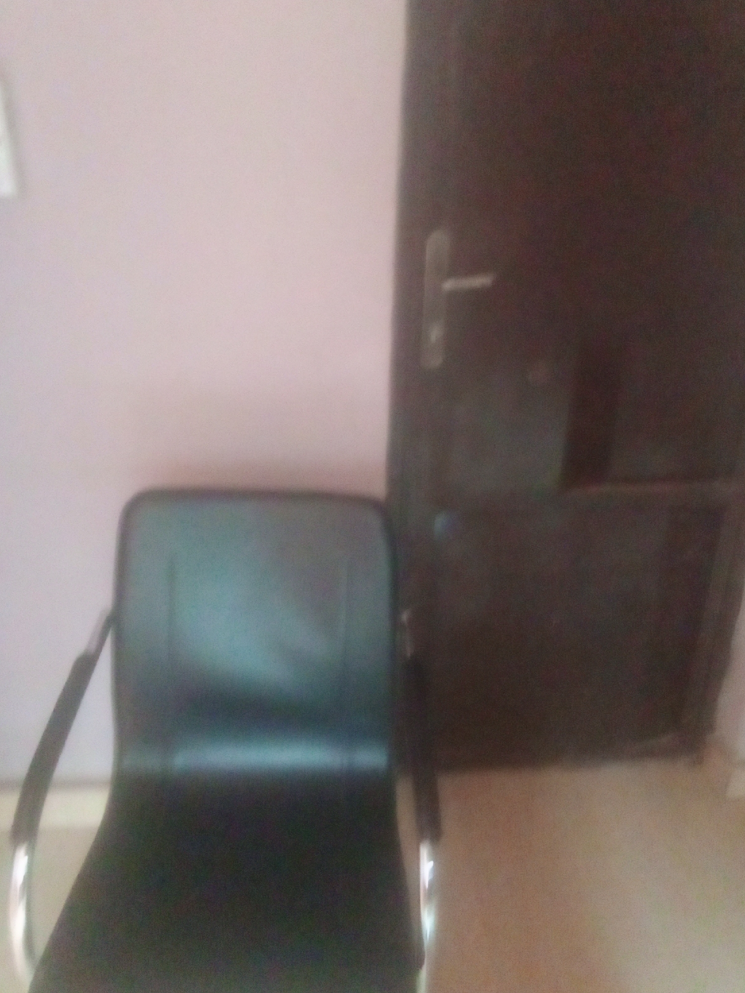 The media file depicts an indoor scene featuring a black chair positioned in front of a dark brown door. The chair has a metallic frame and appears to be made of leather or a similar material. The door has a silver handle. The background wall is a light pink color. The overall lighting suggests an indoor setting, possibly during the daytime. The image is slightly out of focus, making it difficult to discern finer details. No people are visible in the media file, and no specific activity or event is taking place. There is no visible text. The location context is Jalingo, Nigeria.
