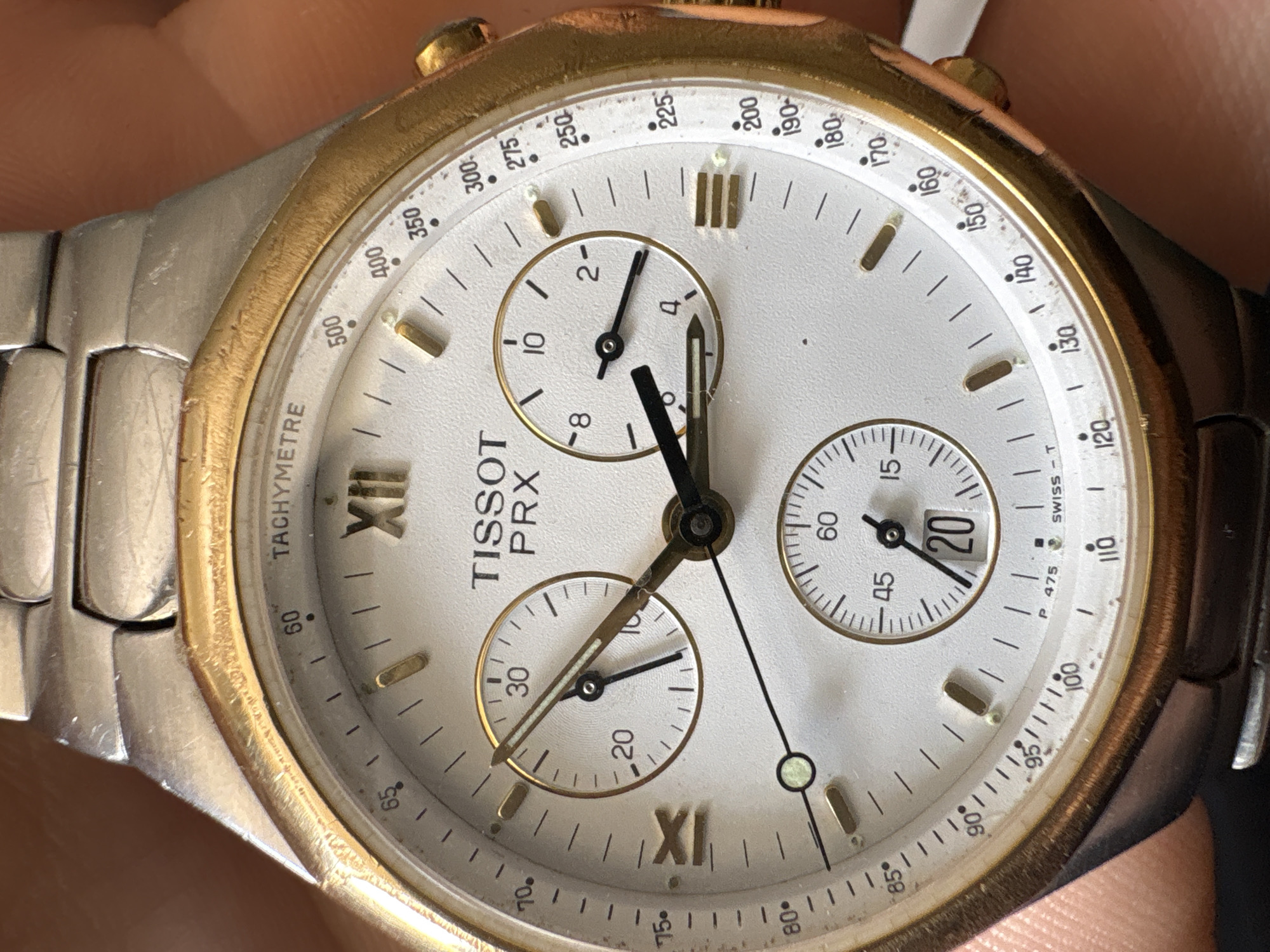 The image shows a close-up of a Tissot PRX chronograph watch. The watch has a silver and gold-tone case and bracelet. The dial is white with gold-tone indices and hands. The subdials display seconds, minutes, and hours. The date window is located at 4 o'clock. The tachymeter scale is on the outer ring of the dial. The watch appears to be in used condition with some wear and tear visible on the case and bracelet. The image does not depict any location in Sofia, Bulgaria, or any other specific location. There are no people visible in the image.