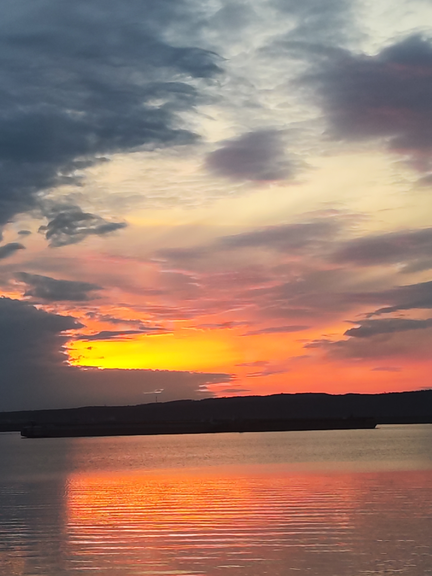This vertical shot captures a vivid sunset over a wide body of water in Chaikovsky, Russia. The sky is a dramatic canvas of colors, with the lowest portion dominated by an intense band of bright yellow and orange, suggesting the sun has just dipped below the horizon or is partially obscured by clouds. Above this fiery glow, the sky transitions through shades of red and pink, blending into lighter orange and pale yellow hues further up. Dark, heavy grey clouds are scattered across the upper and middle portions of the sky, providing a stark contrast to the brilliant sunset colors, with some appearing to be illuminated from behind.
In the midground, a dark, silhouetted landmass, likely a distant shoreline or hills, stretches horizontally across the image, separating the water from the vibrant sky. A long, dark vessel, possibly a barge or cargo ship, is visible on the water, also silhouetted against the reflective surface. The water itself is calm with gentle ripples, acting as a mirror that spectacularly reflects the fiery colors of the sunset, creating a shimmering path of orange and red directly below the brightest part of the sky. The reflection intensifies the warmth of the scene.
The overall atmosphere is serene and awe-inspiring, highlighting the natural beauty of the sunset. There are no people visible, nor is there any discernible text in the image. The time of day is clearly evening, during or immediately after sunset.