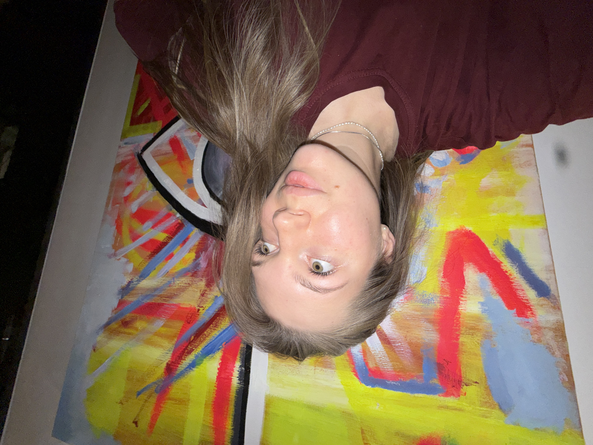 A woman with long, light-brown hair is lying upside down with her head resting on a painting. She is wearing a maroon-colored shirt and a silver necklace. The painting has a yellow background with broad strokes of red, blue, and white paint. There is also a section of the painting with a black circle surrounded by a white square. The picture is taken indoors, and the location is impossible to determine from the image.