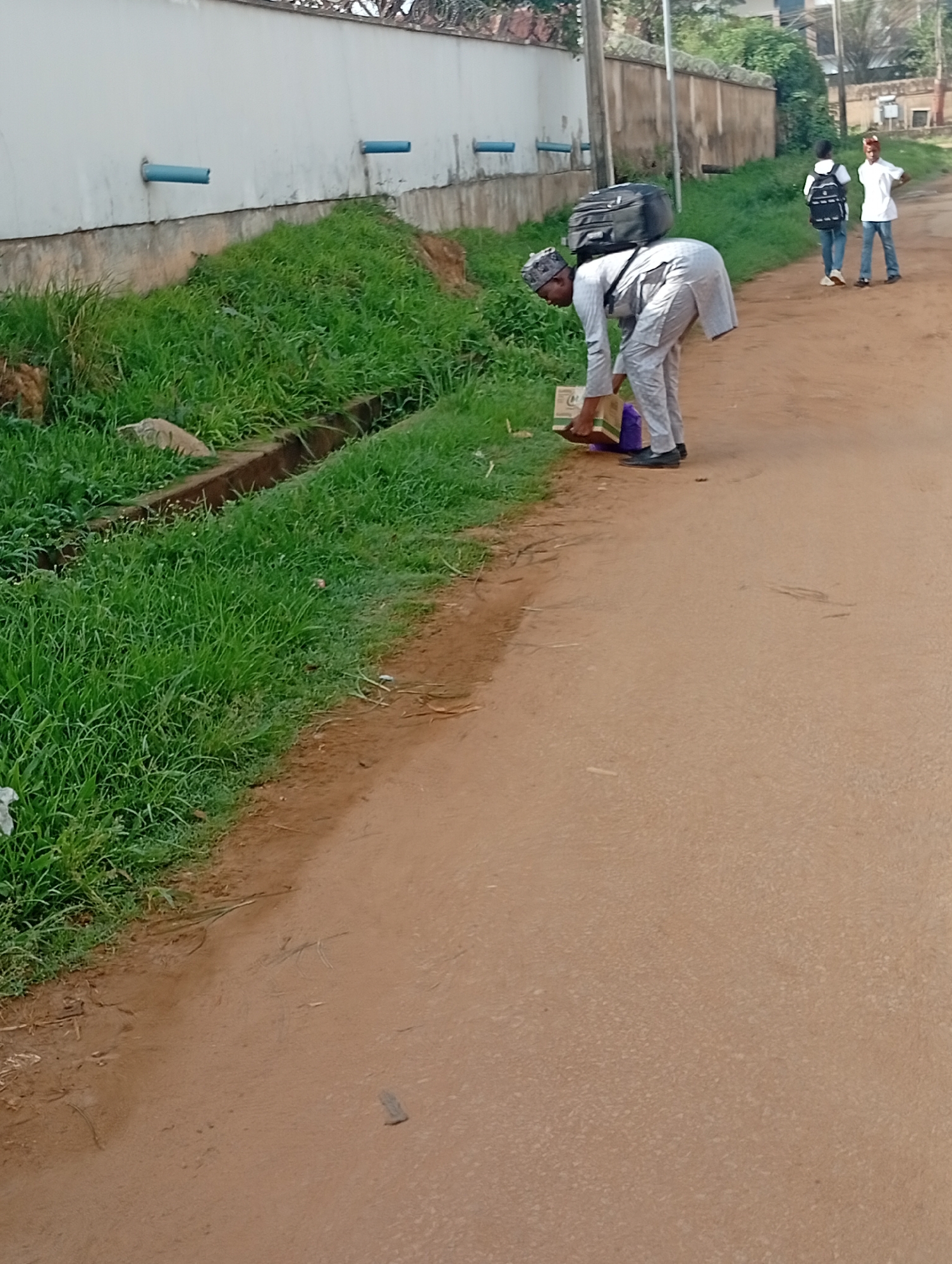 In Rafin Par, Nigeria, a man wearing a traditional grey outfit and cap, with a black backpack strapped to his back, is bending down to pick up items from the ground beside a grassy area. He is holding a yellow and white bag in his left hand. To his right, two young people walk away; one wearing a white t-shirt, jeans and a red cap, and another in a white t-shirt, jeans, and a black backpack. A concrete wall with blue pipes runs along the left side of the image, bordered by a drainage ditch and overgrown grass. The scene is set on a dirt road.