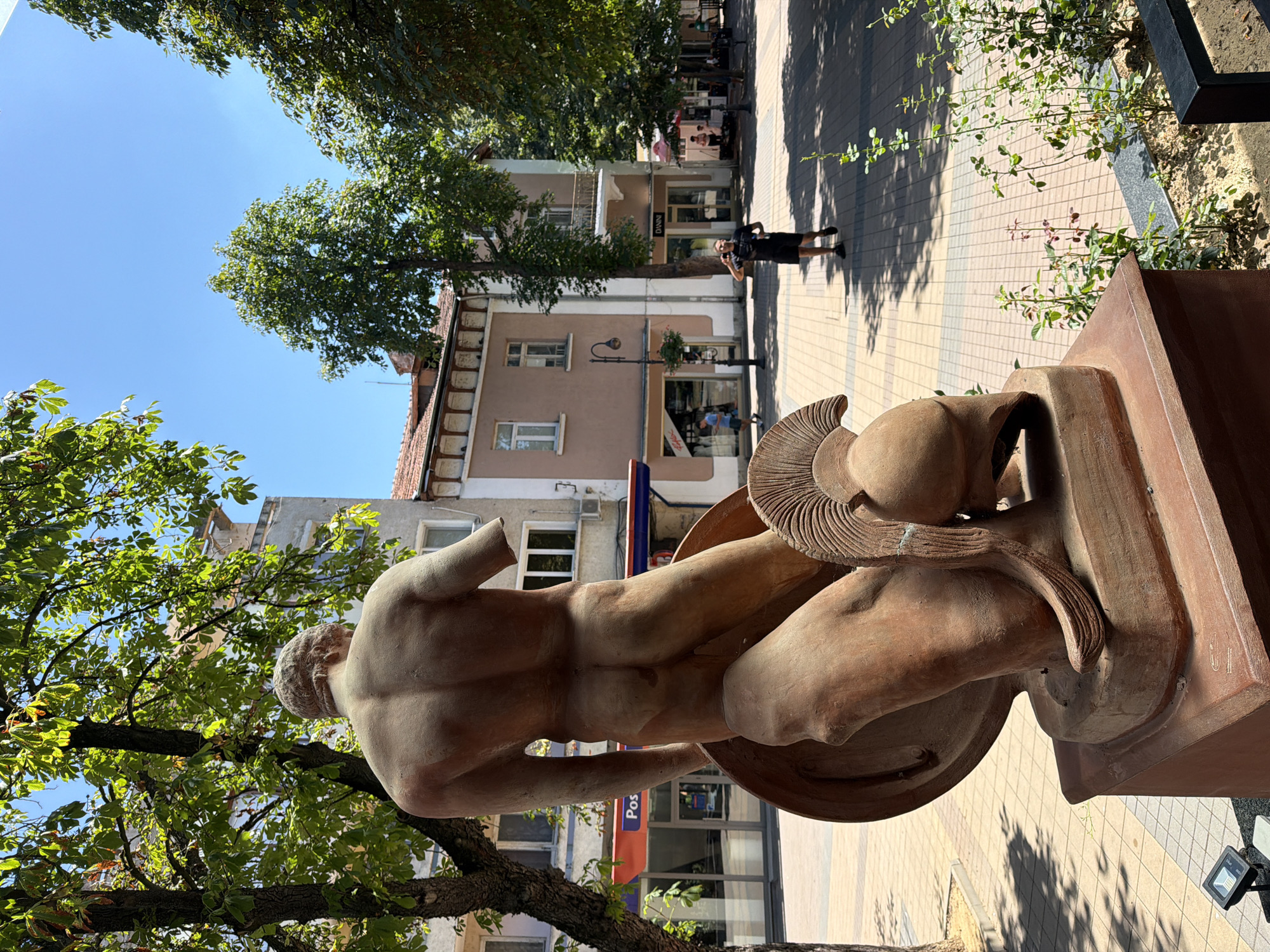 An outdoor scene depicts a statue of a warrior in the city of Lovech, Bulgaria. The statue, made of a reddish-brown material, shows a muscular male figure from the back, with one arm missing. The warrior is depicted holding a shield and wearing a helmet. The statue is situated on a pedestal in a public area with a paved walkway and trees. In the background, there are buildings with windows and balconies, and a few people are visible walking on the street. The sky is clear and blue, indicating a sunny day.