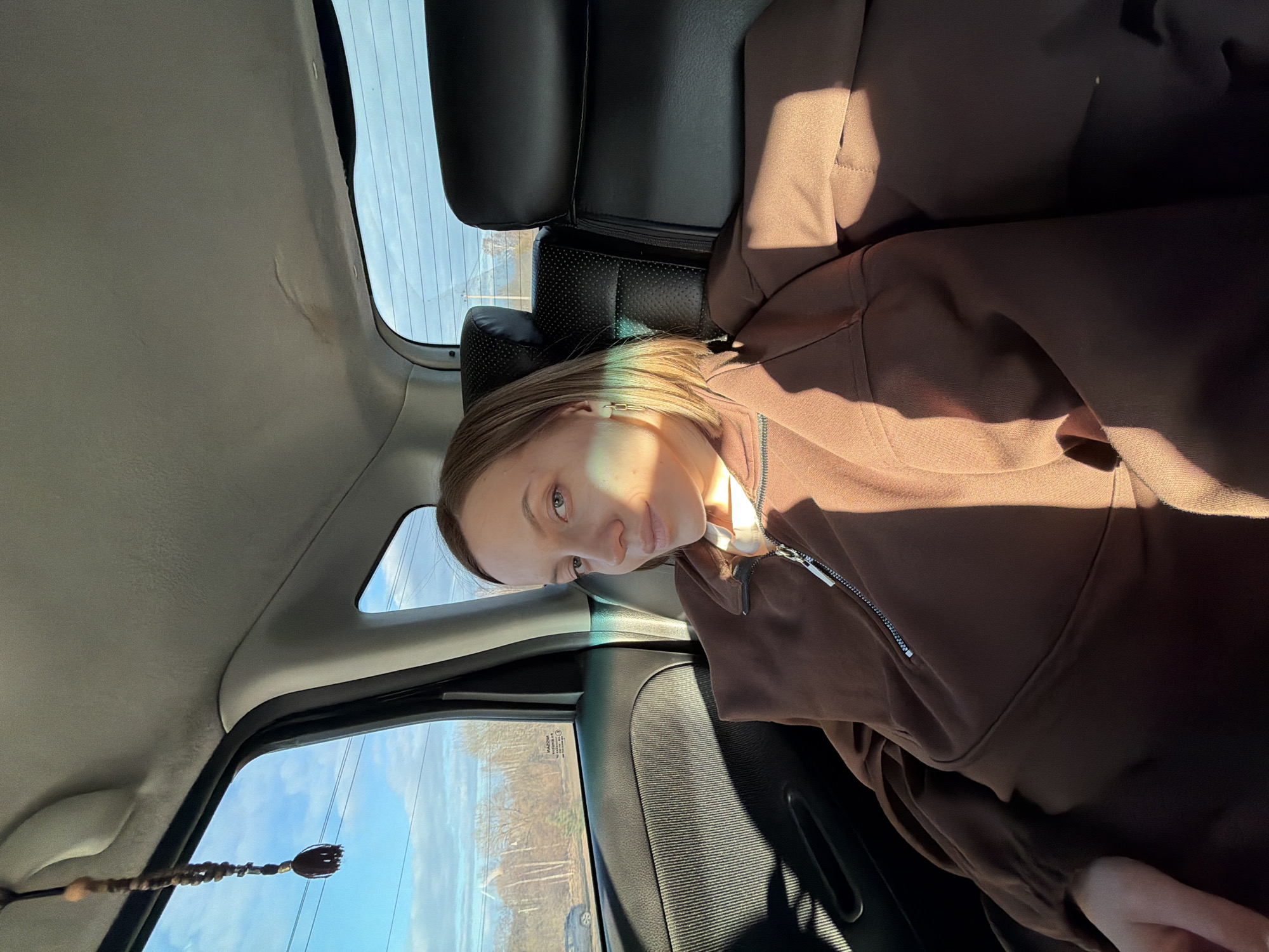 A young, fair-skinned person with light brown hair, wearing a brown zip-up hoodie, is positioned in the backseat of a car. The car's interior is visible, including the leather seats and the window frame. Through the window, a glimpse of the outdoors is apparent, featuring trees and power lines, suggesting a rural or suburban landscape. The picture suggests the car is moving. The person is looking toward the camera. The location is Minino, Russia.