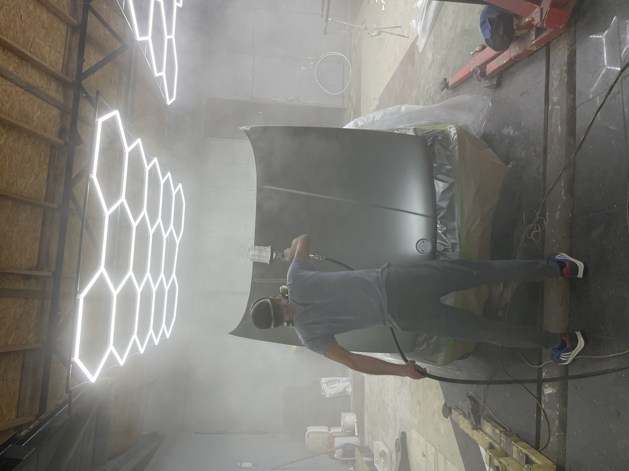 A person is spray painting a car hood in a workshop. The person is wearing a mask, a gray t-shirt, and dark pants. They are holding a spray gun and are positioned to spray the hood, which is propped up and covered in plastic sheeting. The workshop is dimly lit, with bright, hexagonal LED lights on the left side of the frame. The floor appears to be concrete and there is dust or mist in the air from the painting process. In the background, there is a red car lift, various containers, and equipment. The overall impression is of an active workspace where automotive painting is taking place. The image is oriented vertically.