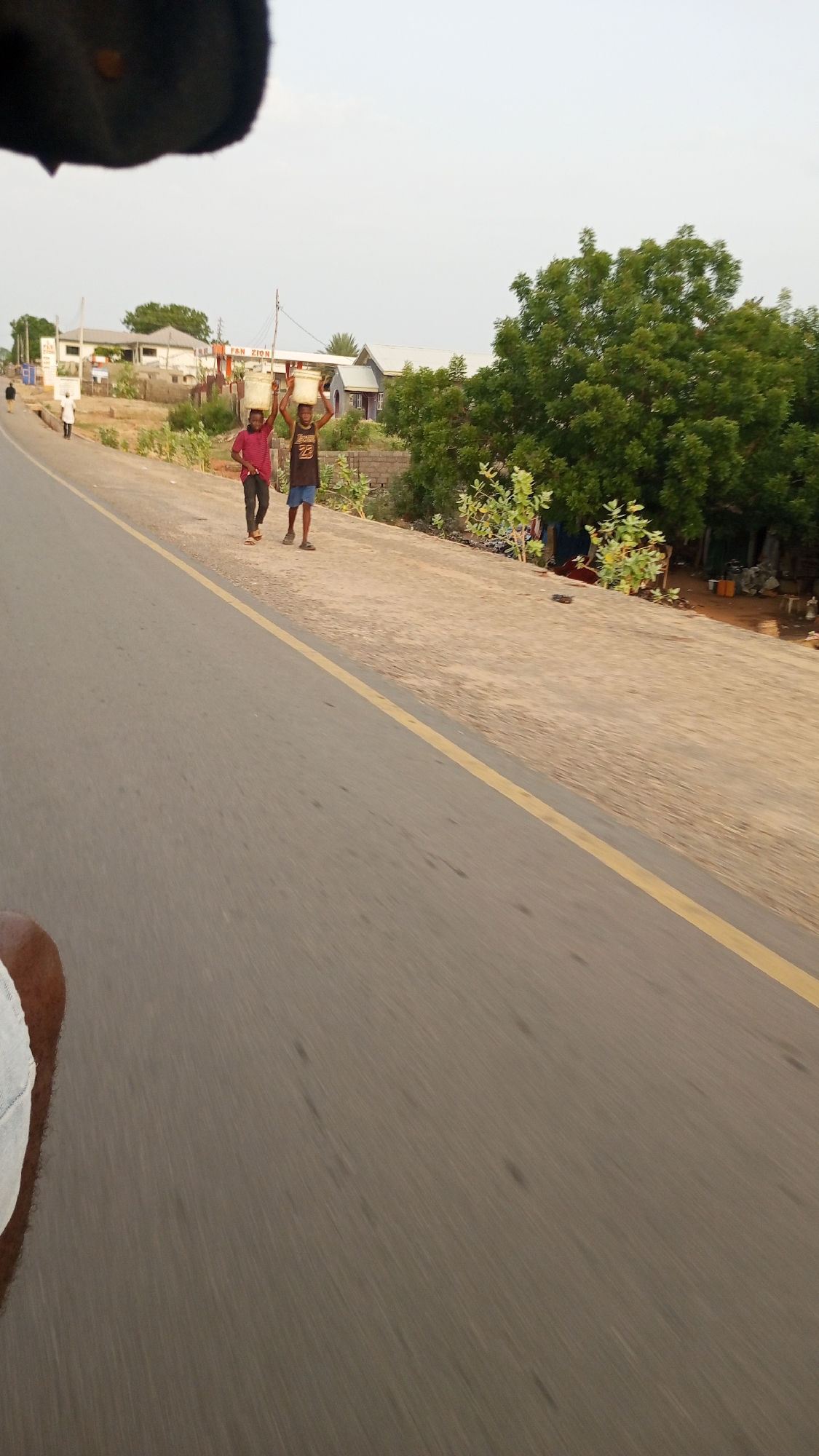 The image shows a road in Zango-ndaji, Nigeria. Two people walk on the shoulder carrying containers on their heads. To the right of the shoulder, there are trees and vegetation. Behind the people and vegetation, there are buildings, including one that appears to be named "FAN ZION". A few other people can be seen walking further up the road. The camera is positioned from a moving vehicle on the road, and a yellow line marks the road's shoulder.