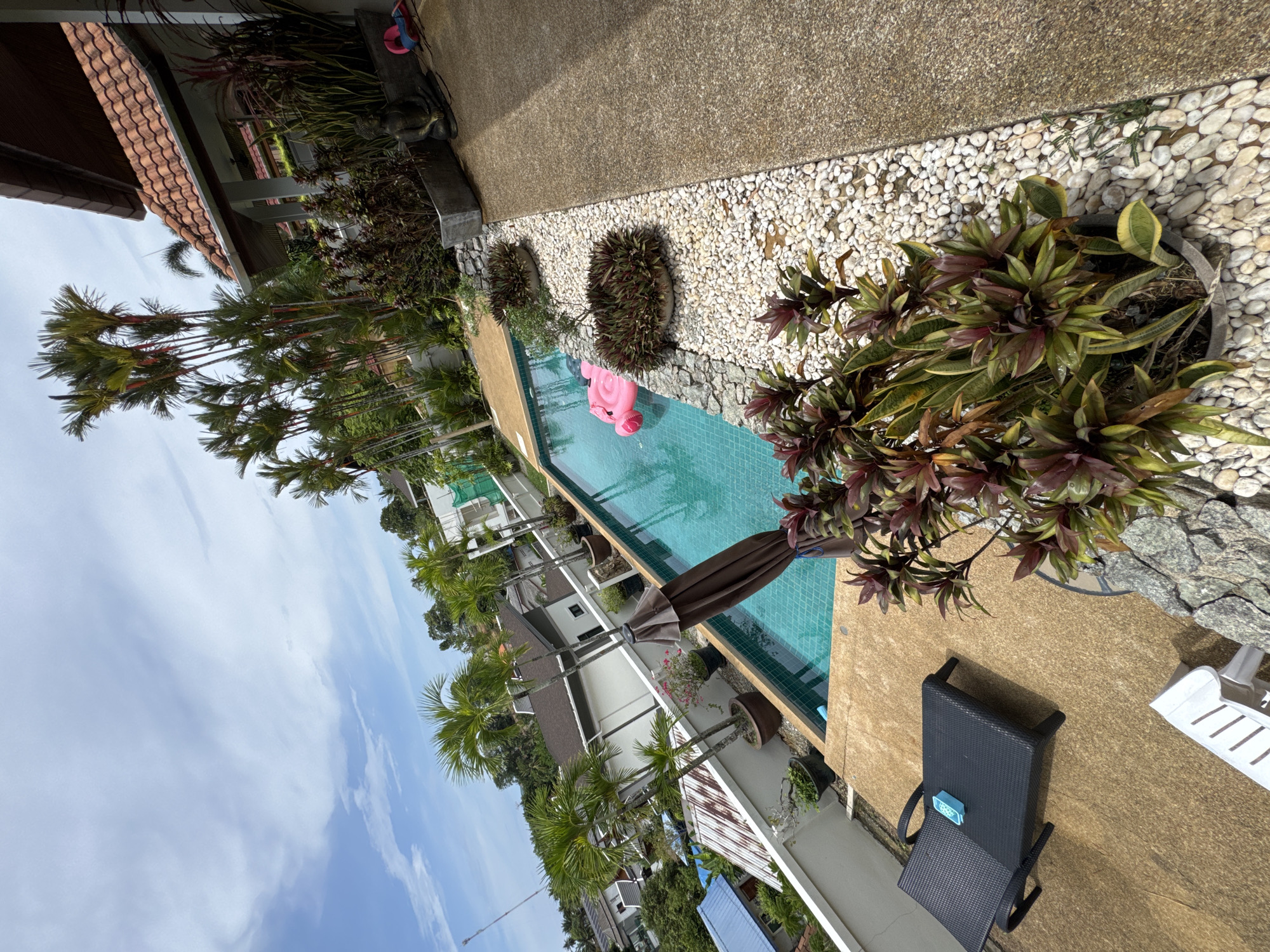 Here's a description of the image:
The image depicts a swimming pool area in what appears to be a tropical resort. The pool is lined with small white rocks, with various potted plants along the edge. A pink inflatable flamingo is floating in the pool. A dark-colored lounge chair with a blue item on it is placed on a light brown surface next to the pool. In the background, there are residential buildings and palm trees. The sky is blue with some clouds. Based on the location, Rawai, Thailand, this scene suggests a private residence or resort villa.