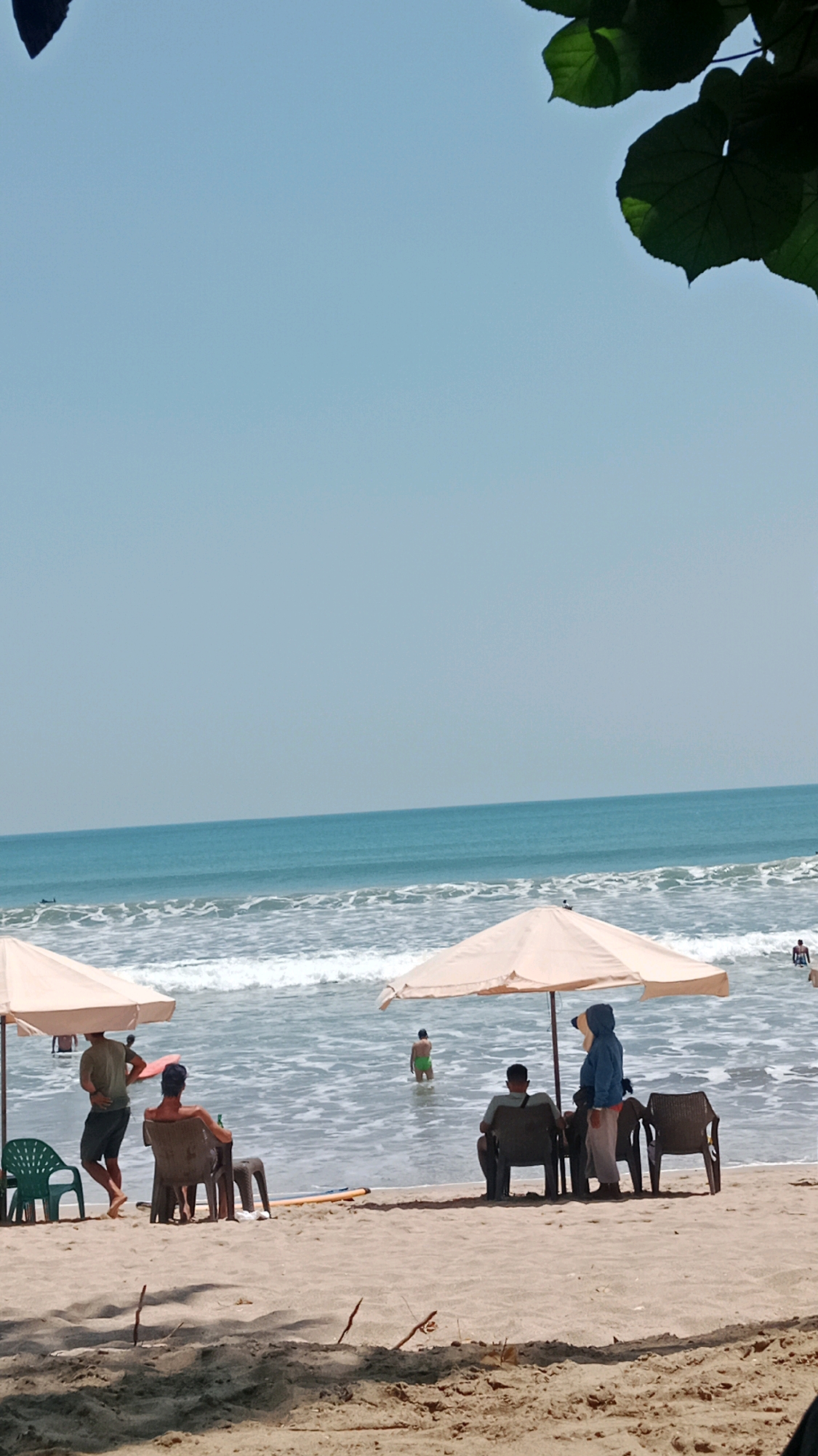The image depicts a sunny day at Kuta Beach, Bali, Indonesia. In the foreground, the sandy beach is visible with a few pieces of driftwood. Several beach chairs are set up under two light-colored umbrellas. On the left, under one umbrella, a man wearing a green t-shirt and dark shorts stands, while another man sits in a chair looking out at the ocean. To the right, under the second umbrella, two people are seated. One person, wearing a blue hooded jacket and light-colored pants, stands next to a seated person. In the background, the ocean has moderate waves, and several people are in the water. One person is visible standing in the shallow water wearing green shorts, and another person is further out in the water. The sky is clear and blue.