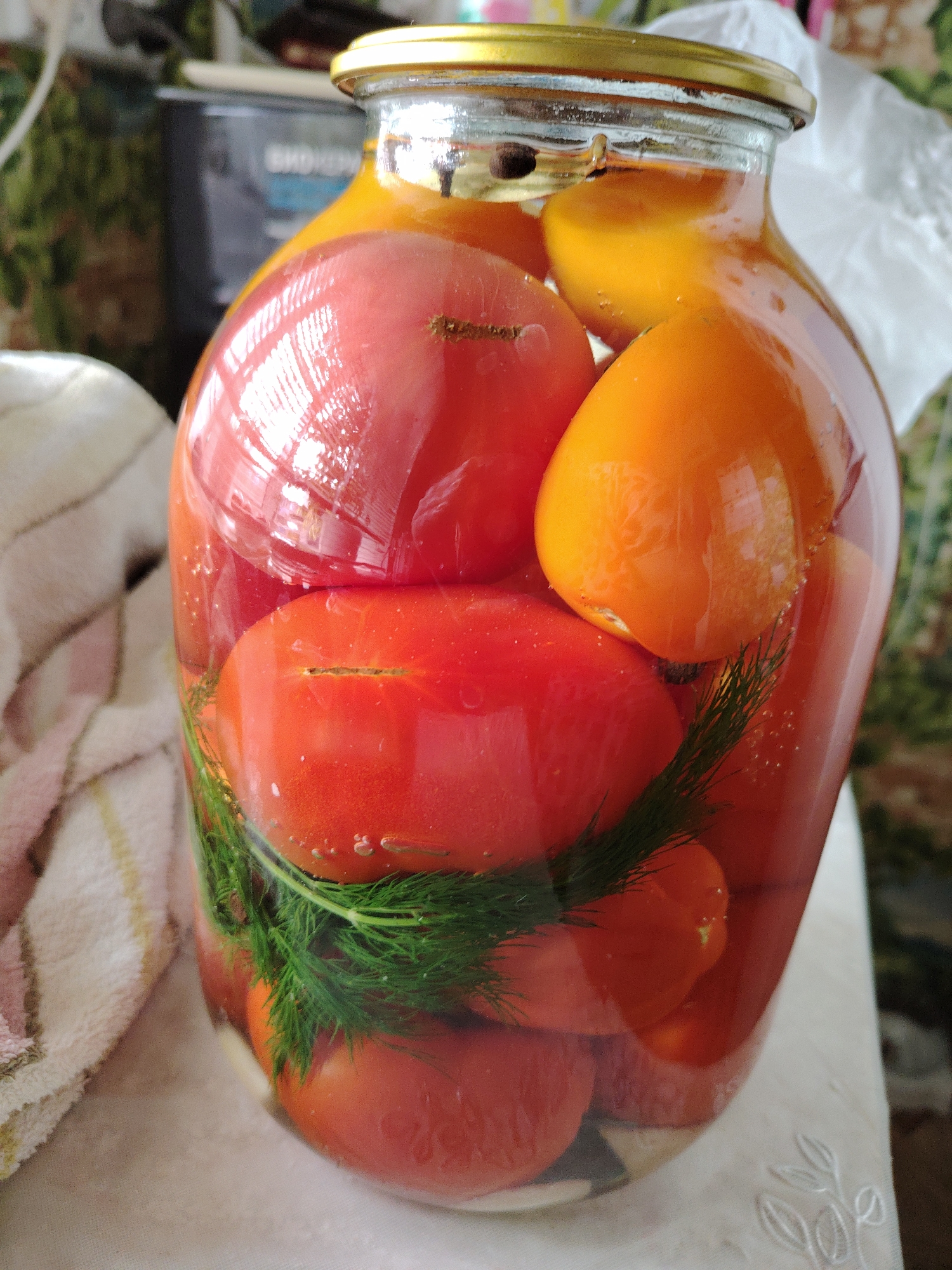 A close-up, vertical shot focuses on a glass jar filled with preserved red and yellow tomatoes, as well as dill. The jar has a gold-colored lid. Bubbles are visible within the brine surrounding the tomatoes. The jar is placed on a white, textured surface, possibly a tablecloth, which has a subtle embroidered pattern of leaves on the right side. To the left of the jar, a striped cloth with pink, white, and yellow stripes is visible. In the background, a blurred image of a dark appliance, likely a kitchen appliance, and a patterned curtain or wallpaper can be seen. The lighting in the scene creates reflections on the glass jar and the tomatoes. The overall setting suggests a domestic kitchen environment.