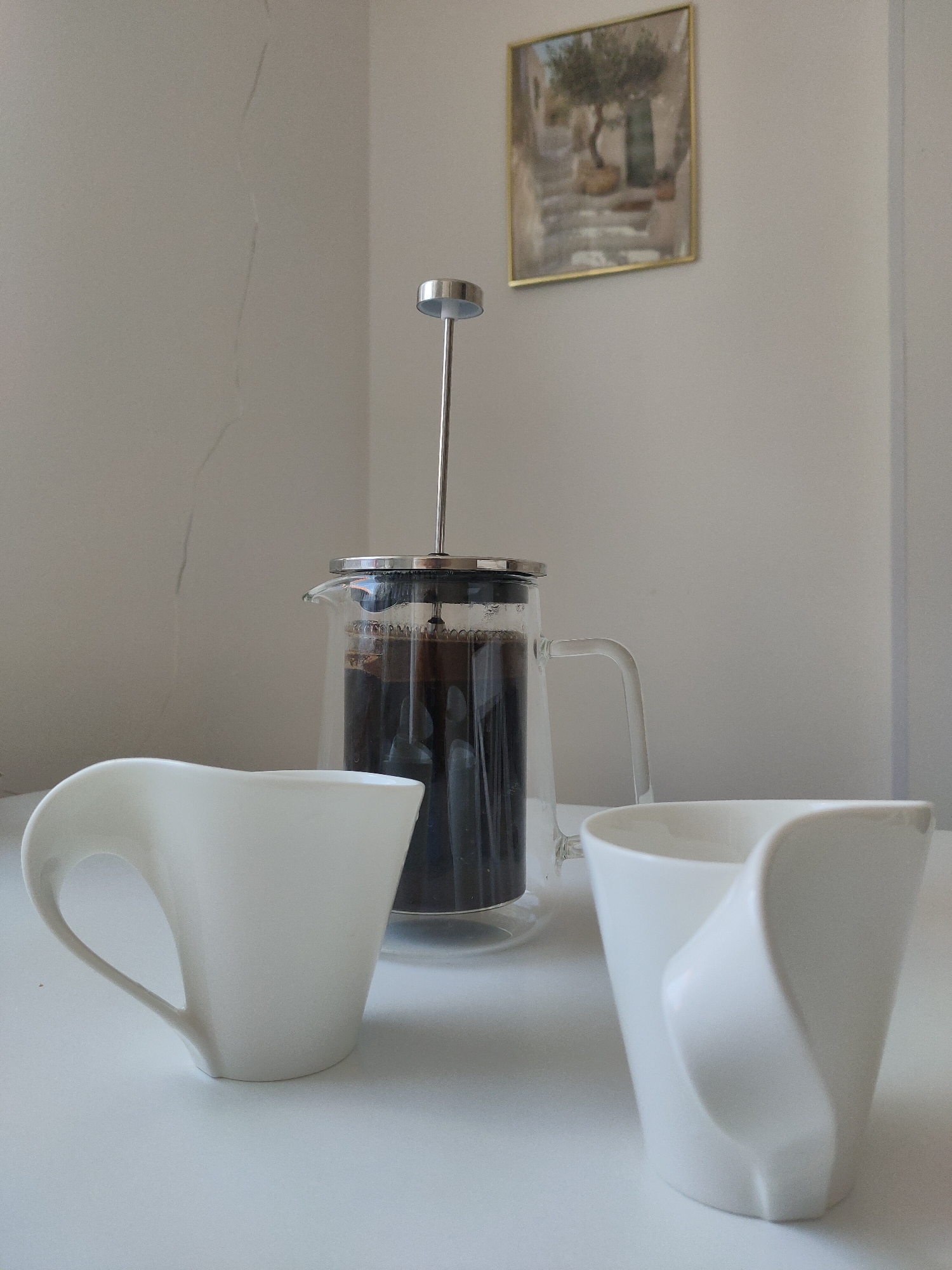 The image shows a table setting with a French press coffee maker and two white ceramic mugs. The French press is made of glass and metal, and it contains dark coffee. The mugs have a modern, sculptural design. In the background, a framed picture of a Mediterranean-style scene with an olive tree hangs on a light-colored wall. The wall has a faint crack running vertically down it. The scene appears to be indoors, possibly in a home or a cafe. There are no people visible in the image. The overall impression is one of a peaceful, quiet moment, perhaps a break for coffee. There is no information that would place this scene in Zrenjanin, Serbia.