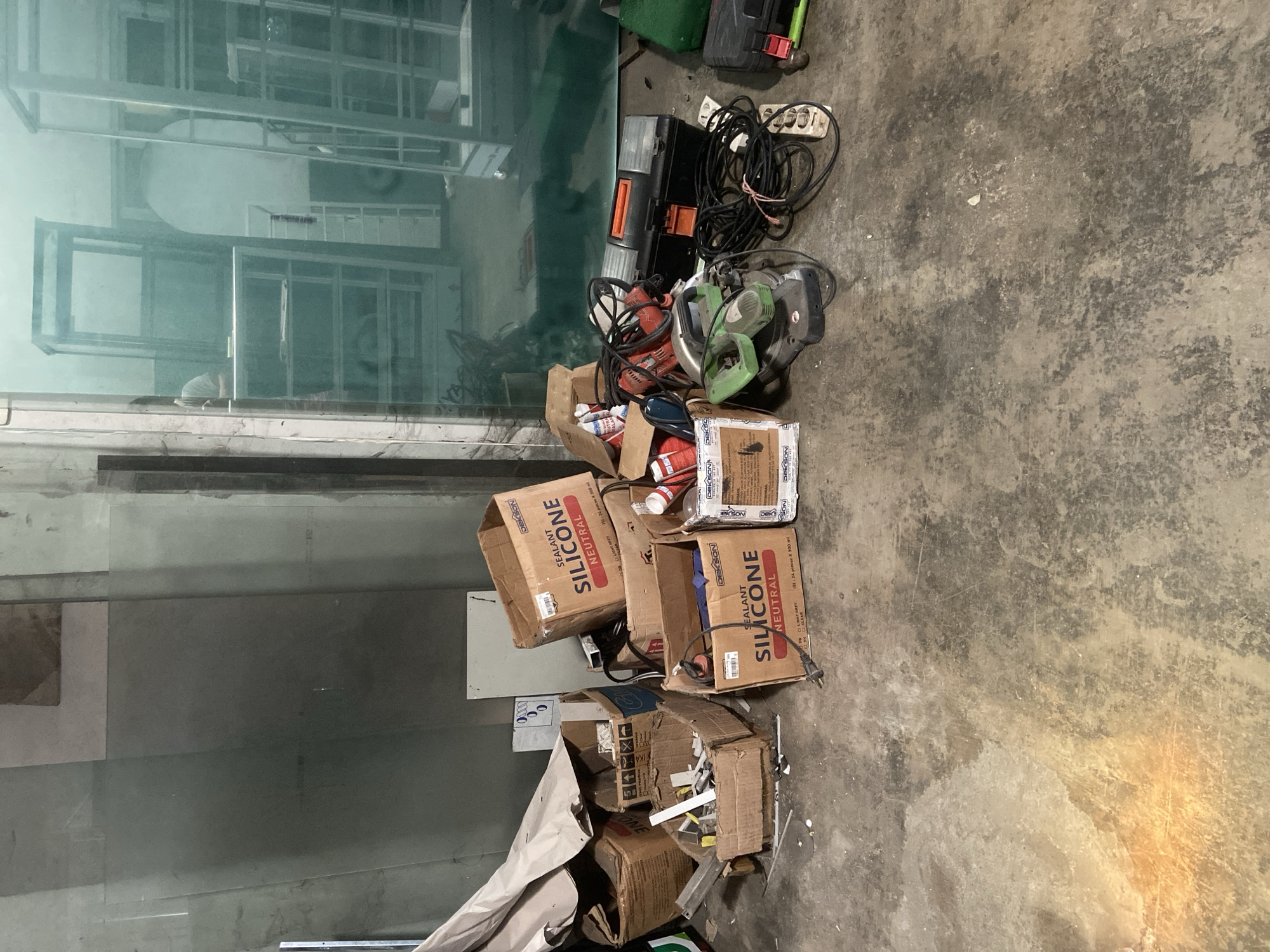 The image depicts an indoor storage area, possibly a workshop or warehouse, in Plosokandang, Indonesia. The scene is cluttered with various construction materials and tools scattered across a concrete floor. Prominently featured are several cardboard boxes, some labeled "DEKSON SILICONE NEUTRAL." Among these boxes are tubes of silicone sealant, tools such as a green and black miter saw, a red power drill, and a black toolbox with orange accents. Electrical cords and an extension cord are also visible, tangled amongst the items. In the background, large glass panels lean against a wall, and metallic shelving units are present, suggesting a retail or display area. The lighting in the scene is somewhat dim, with a warm glow emanating from the lower left corner, possibly from an artificial light source. No people are clearly visible in the immediate foreground or among the items. The overall impression is one of disarray and the storage of building or renovation supplies.