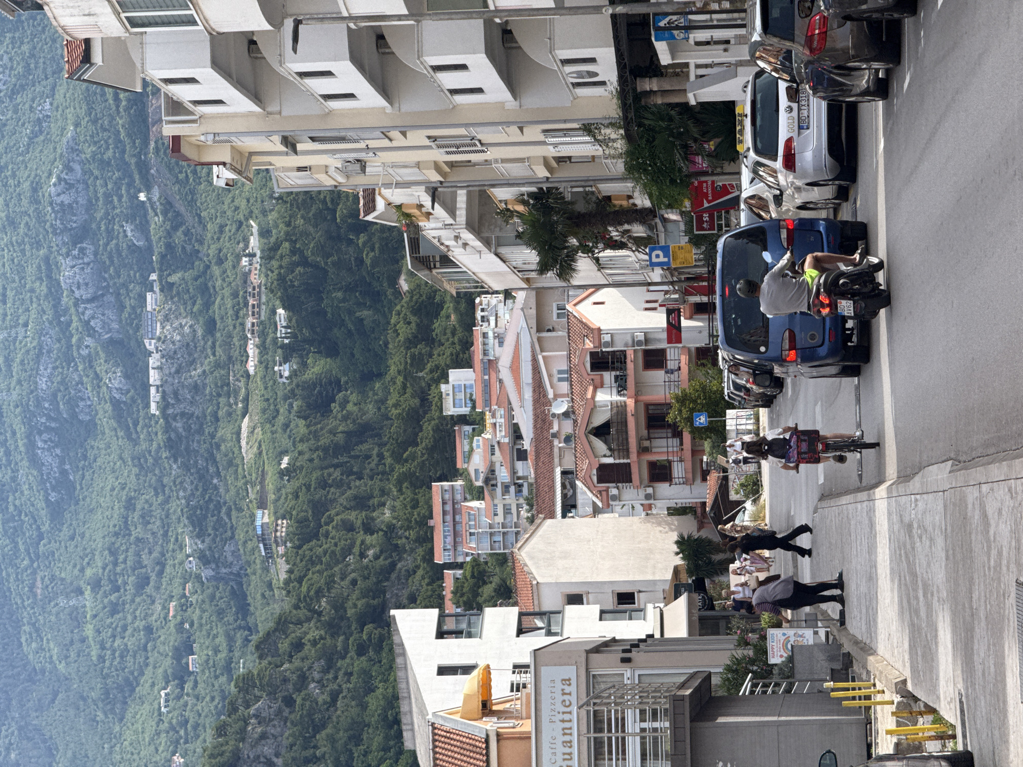 This image depicts a bustling street scene in Budva, Montenegro, under bright daylight conditions. The perspective is slightly elevated, looking down a street flanked by buildings on the left and parked vehicles on the right, with a vast forested mountain landscape in the background.
On the right side of the image, a paved street features several parked cars. A blue car is prominent, behind which a person on a scooter, wearing a helmet and shorts, appears to be in motion or waiting. Among the parked vehicles is a silver Volkswagen Golf taxi, identifiable by the "TAXI GOLD" sign on its roof and "BD TX318" on its license plate. Another car has the license plate "BD AV62". On the adjacent sidewalk, several pedestrians are visible, including one person pushing a bicycle equipped with a basket. Yellow bollards or barriers line the edge of the pavement.
To the left, a succession of multi-story buildings lines the street, displaying a mix of modern apartment blocks with balconies and structures with traditional tiled roofs. One building prominently features a sign that reads "Caffe - Pizzeria Quantiera". Further down the street, smaller signs indicate "HAPPY KIDS" and "ATM BANKOM". Palm trees are scattered among the buildings, enhancing the coastal ambiance.
The background is dominated by a sweeping view of heavily forested mountains, with various buildings and structures nestled within their green slopes, extending into the distance under a clear, bright sky. The scene portrays typical daily life in a vibrant, sunny coastal town.