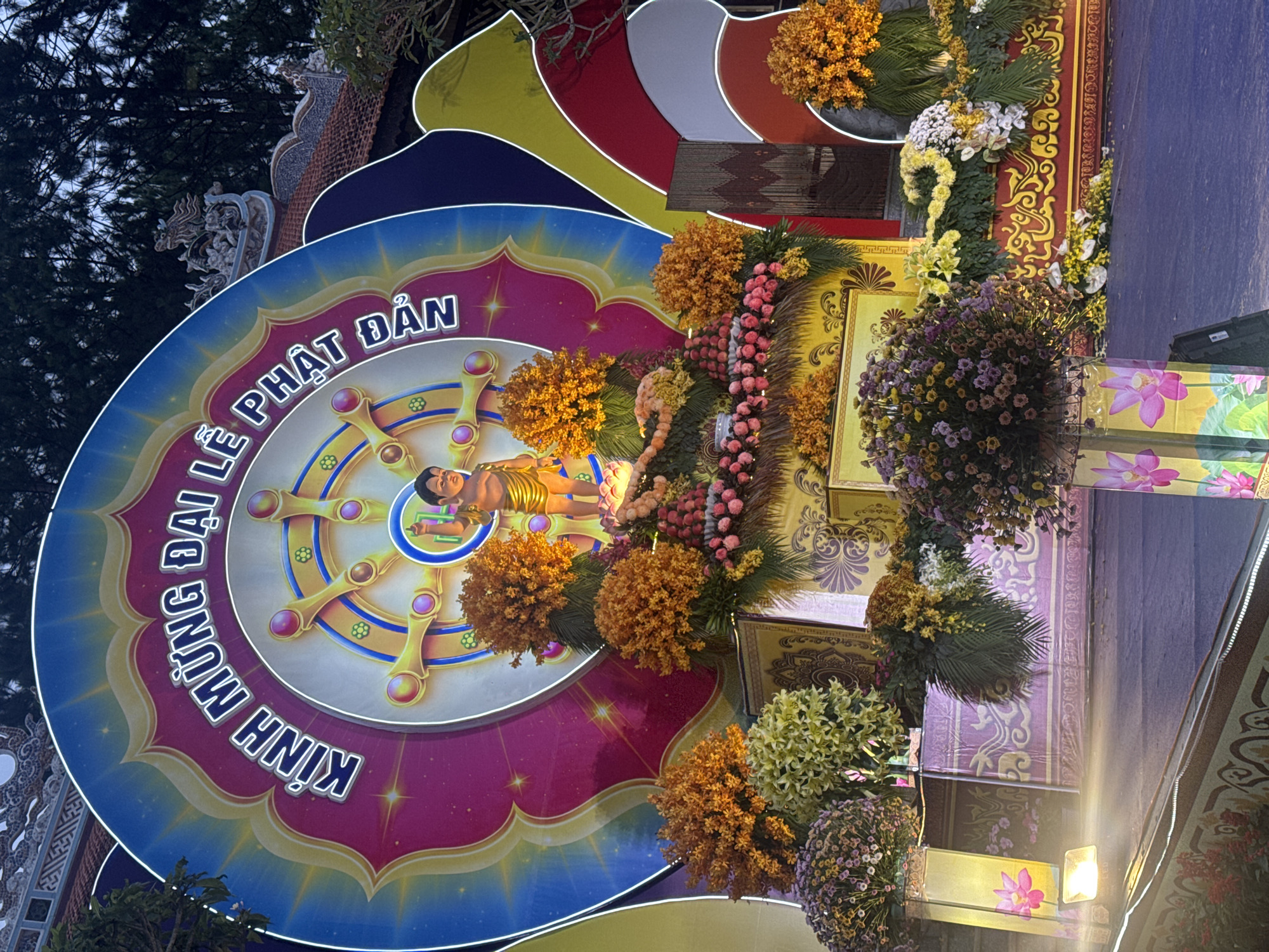 The image shows a festive Buddhist display with a colorful backdrop, likely erected for a celebration. Centrally positioned is a large, circular sign featuring the Dharmachakra (Wheel of Dharma), a key Buddhist symbol. A golden, reclining figure of Buddha is placed in front of the wheel. The sign has Vietnamese text around it, and is surrounded by colorful concentric circles in shades of blue, red, yellow, and white.
The display is decorated with numerous flower arrangements in various colors including orange, yellow, and purple. The backdrops consist of ornate panels featuring traditional Vietnamese designs.
Given the presence of Vietnamese text on the sign, and based on visual evidence from similar scenes online, it is probable that the image was taken in Da Lat, Vietnam. The image suggests an outdoor celebration, possibly a religious holiday like Vesak, observed by Buddhists worldwide. The picture was taken from a low angle.