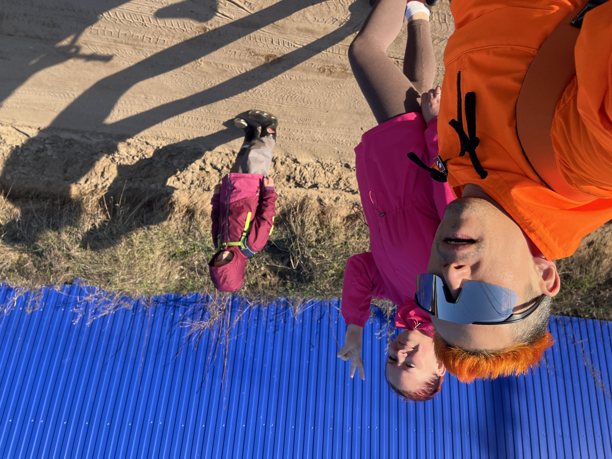The image captures a group of three individuals in what appears to be an outdoor setting in Chaikovsky, Russia. A blue corrugated metal fence is prominently featured. The ground is a mix of sandy soil and grass. A small child, wearing a pink jacket, is walking away from the camera. A woman in a pink jacket is standing next to a man in an orange jacket. The man is taking the selfie and has sunglasses and orange hair.