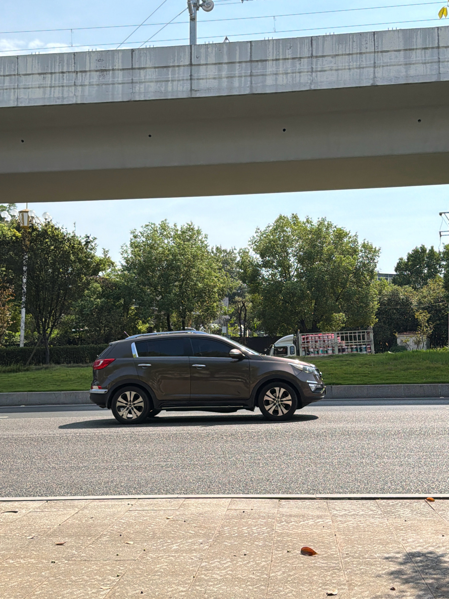 An eye-level, medium shot captures a brown SUV driving on a paved road in the city of Jiangjin, China. The SUV is positioned in the right side of the frame, facing left. It is a bright, sunny day with a clear blue sky and some clouds visible. In the background, a large elevated concrete structure, likely a bridge or overpass, dominates the upper portion of the image. Below this structure, a line of lush green trees and some bushes stretches across the scene. A white truck carrying several large containers is partially visible behind a metal fence on the right side of the road, surrounded by grass. The road surface is asphalt with a textured appearance. In the foreground, a paved walkway with square stone tiles is visible, with a few fallen leaves scattered on its surface. The overall environment suggests an urban setting with green spaces and infrastructure.