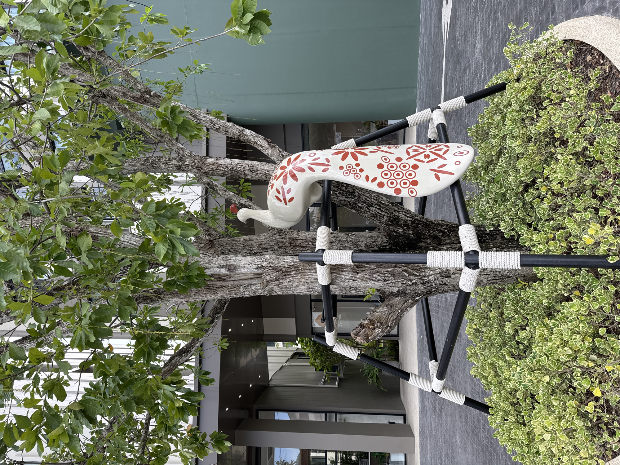 A white, ceramic peacock statue with red floral and circular patterns is mounted on a black metal tree support structure around the base of a mature tree. The tree has textured bark and green leaves. The statue is positioned on the right side of the tree trunk. The support structure consists of several black poles connected at angles and wrapped with white rope. The tree is surrounded by a low-lying green bush with variegated leaves. The ground is paved with dark gray tiles. In the background, a modern building with large windows and gray siding is visible, along with a large, smooth green wall. The scene appears to be an outdoor public space, possibly a garden or plaza, in Kathu, Thailand.