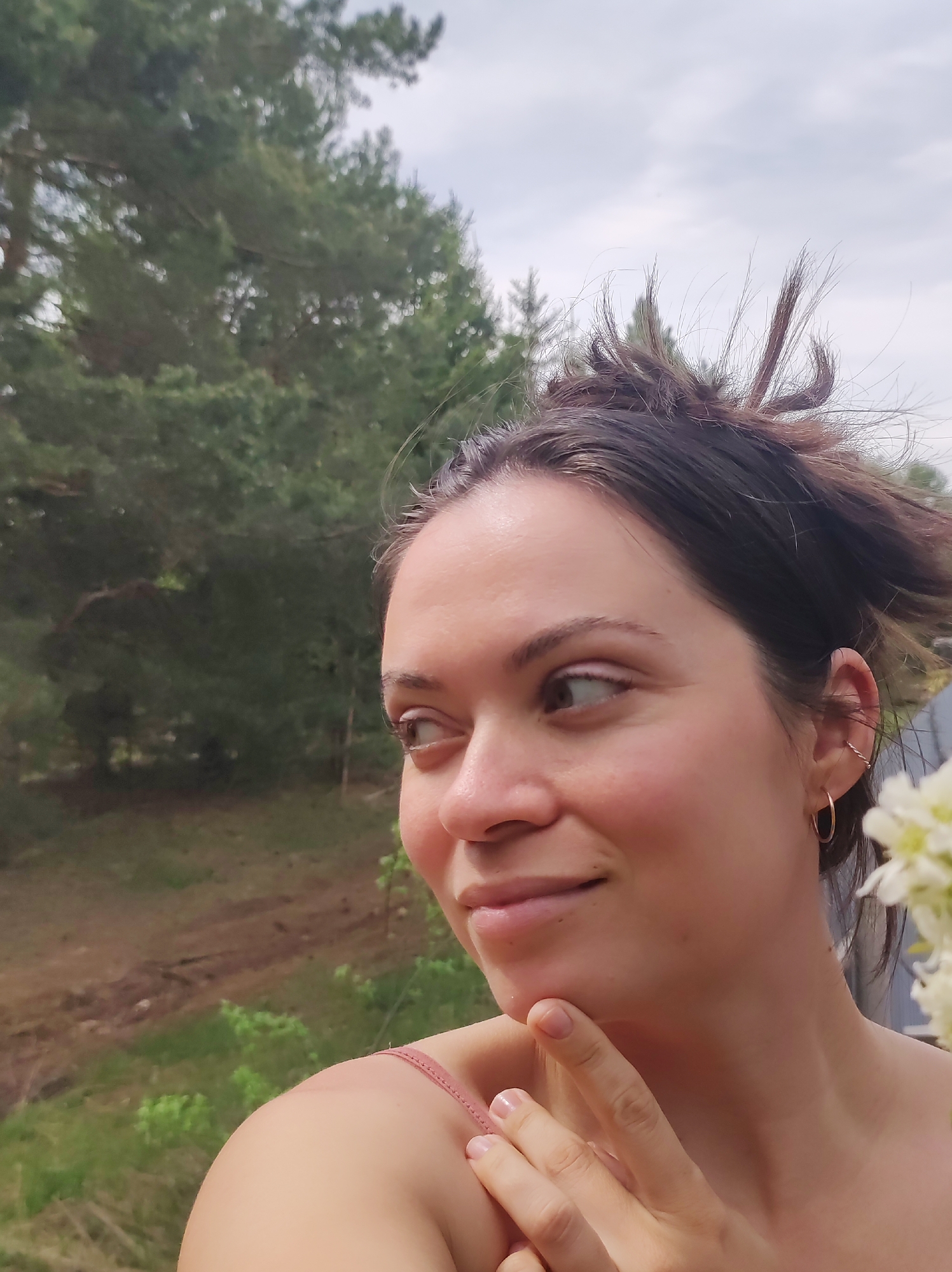 A woman is posing outdoors in a natural setting. The background includes trees and a cloudy sky, suggesting a forest or park. The woman is wearing simple jewelry. She is touching her chin with her hand. There is no evidence of the city of Фоки, Russia.