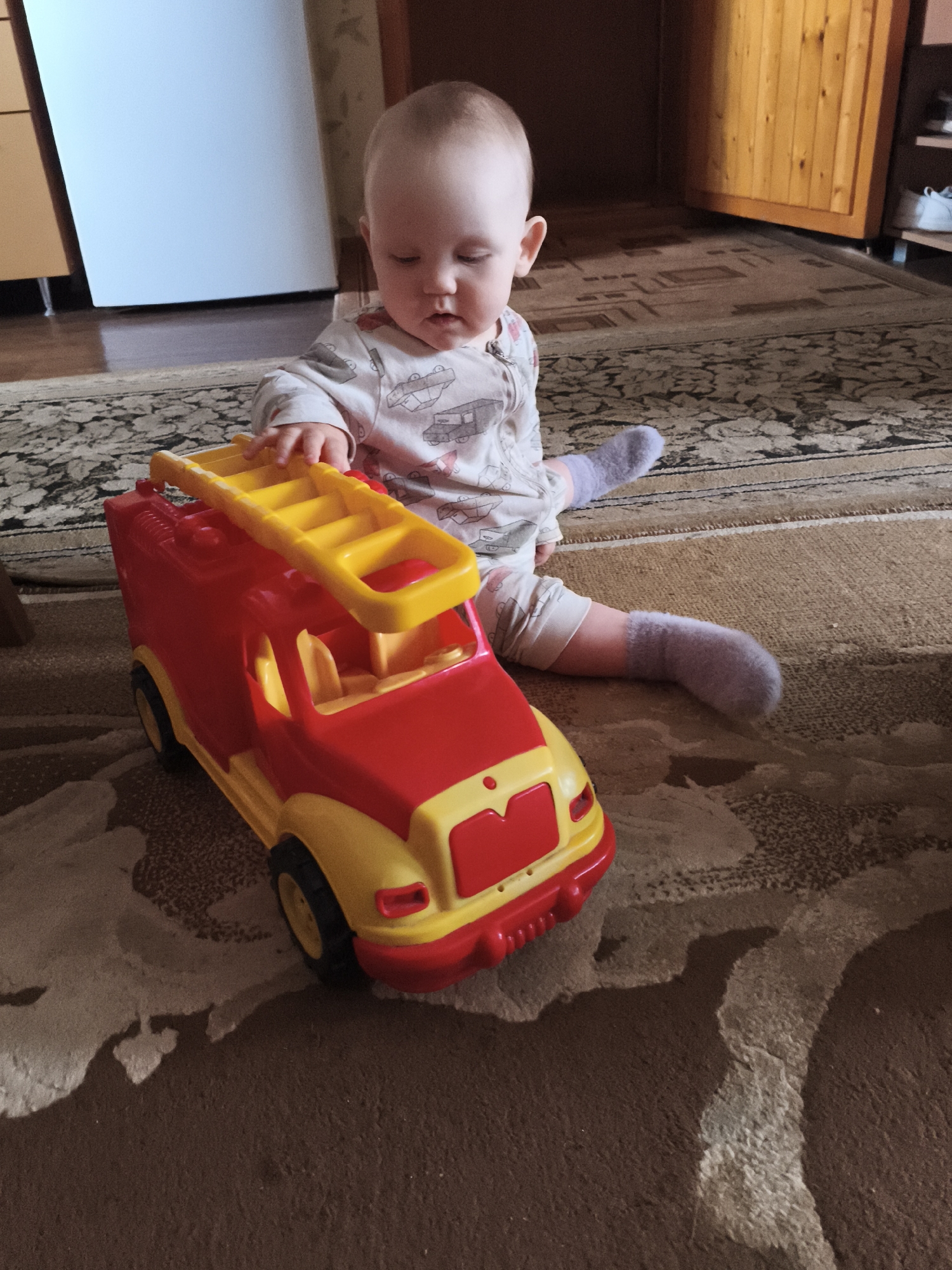 This image captures a single infant seated on a patterned floor rug in an indoor residential environment in Unknown, Unknown. The infant wears a light-colored long-sleeved garment patterned with various vehicles and light purple socks. Their right hand is positioned on the yellow ladder structure of a large, red and yellow plastic toy fire truck, while their head is tilted downwards, indicating focus on the toy. The floor is covered by two distinct rugs: a patterned beige and brown rug beneath the infant and truck, and a darker, more intricately patterned rug further back. In the background, to the left, a portion of a light-colored cabinet or appliance is visible. To the right, a wooden paneled door or cabinet and a white shelving unit holding a pair of light-colored shoes are present. Ambient lighting illuminates the scene, casting subtle shadows.
