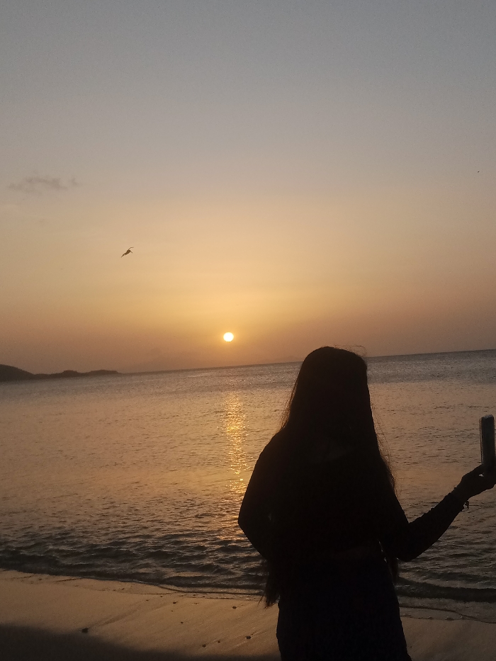 An outdoor scene captured at sunset shows a solitary figure on a sandy beach in Juan Griego, Venezuela. The subject, a person with long hair, is positioned in the lower right quadrant of the frame, presented in silhouette, facing slightly towards the right. They are holding a smartphone vertically in their left hand, extended outward. The sky is characterized by a gradient of pale blue at the top transitioning to vibrant orange and yellow hues near the horizon. The sun, a bright circular orb, is situated above the horizon line, casting a reflective shimmer across the ocean surface. Gentle waves are visible on the water, meeting the sandy shore. A distinct landmass, likely an island or coastline, is silhouetted against the sky on the distant left horizon. One bird is observable in flight in the upper left portion of the sky, with a faint cloud visible below it.