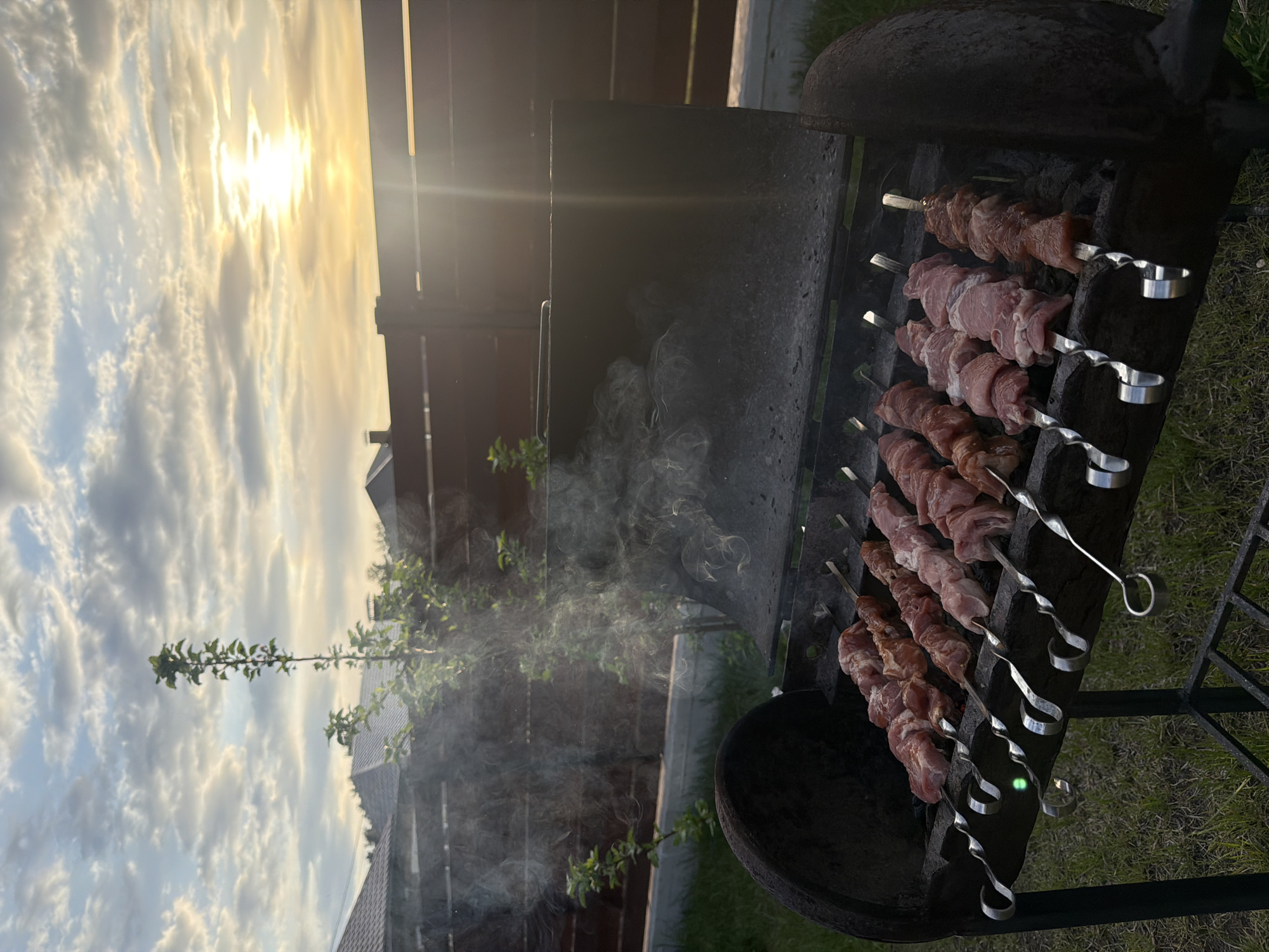 A barbecue grill is located in the city of Chaikovsky, Russia, as inferred from the photo's metadata. The grill contains several skewers with meat, emitting smoke. The background shows a fence, vegetation, and a cloudy sky with the sun shining through. The scene suggests an outdoor barbecue event taking place in a residential area of Chaikovsky.