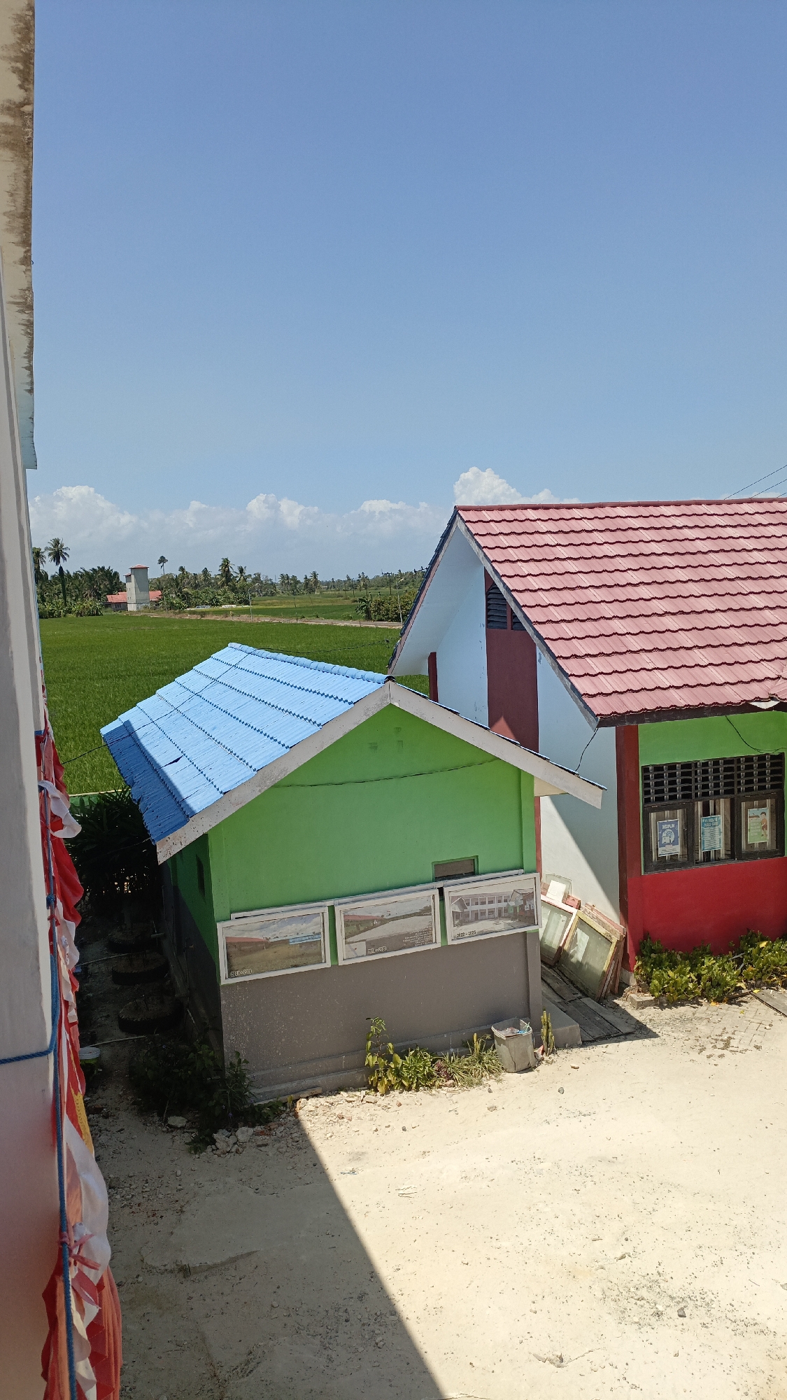 The image depicts a bright, sunny day with a clear blue sky and scattered white clouds, indicating good weather. In the foreground, a section of a white wall with some discoloration and exposed insulation is visible on the left side. Hanging from this wall are red, white, and blue decorative banners, suggesting a celebration or event.
Dominating the midground are two buildings. The smaller building on the left is painted bright green with a light blue, corrugated metal roof. It has a grey base and displays several framed photographs or posters on its wall. Adjacent to it, and slightly further back, is a larger building with a red-tiled roof. The upper part of this building is painted white, with a maroon accent around a small ventilation window. The lower section of this building is painted bright green, with a red base. This building features windows with dark frames and what appear to be posters or notices displayed inside. Several old, wooden-framed windows are leaning against the exterior wall of this building.
The ground in front of the buildings is unpaved, consisting of sandy soil and gravel, with a large shadow cast across it from an unseen object or structure. Scattered vegetation, including small bushes and weeds, grows around the buildings and on the unpaved ground.
In the background, a vast expanse of lush green rice paddies stretches towards the horizon, dotted with scattered palm trees. A tall, cylindrical water tower with a red cap stands in the distance, within the agricultural landscape. The overall scene suggests a rural or semi-rural setting, possibly a school or community area in Saring Sungai Binjai, Indonesia, given the tropical vegetation and the architectural style of the buildings.