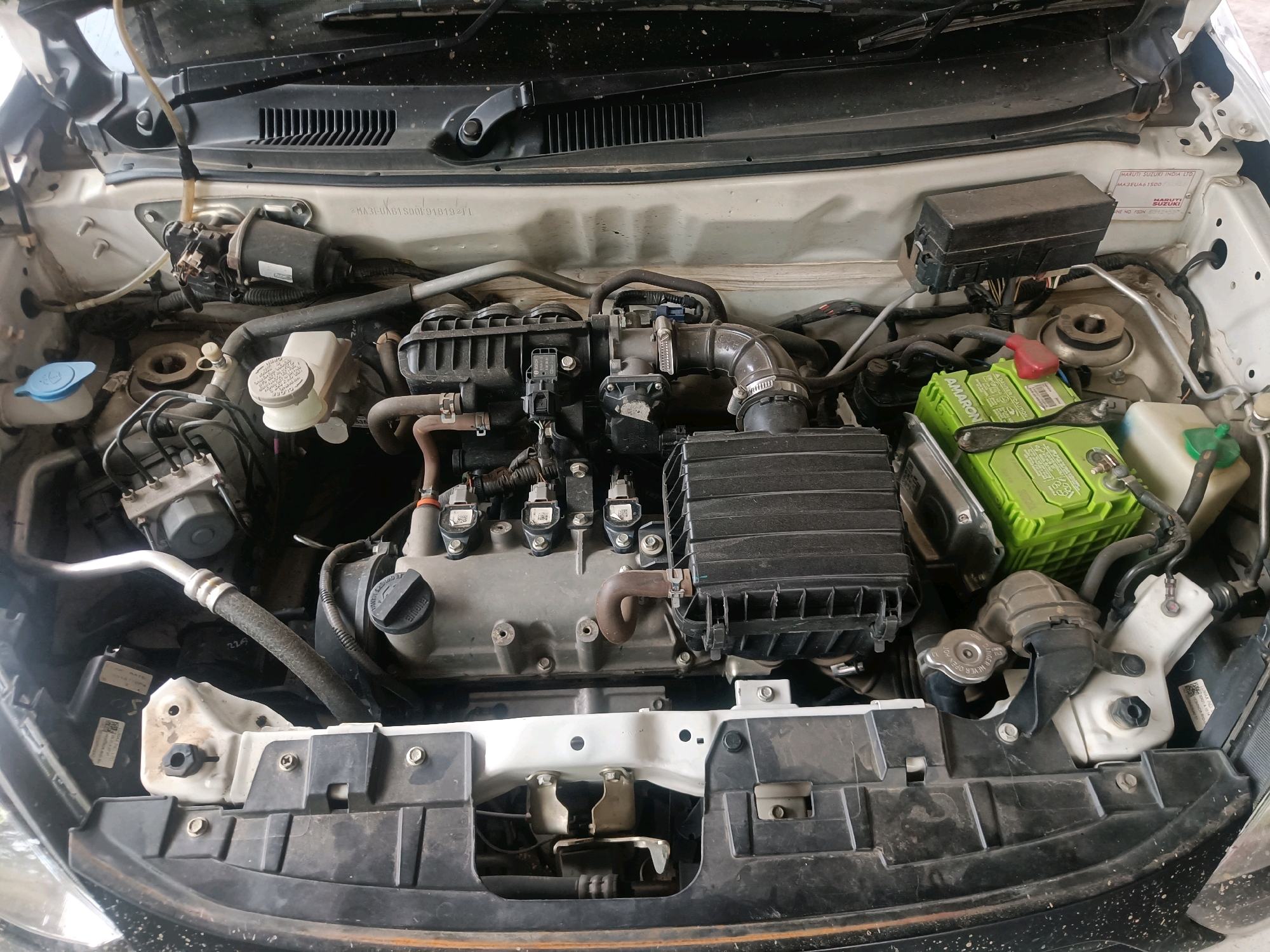 The image shows a close-up, high-angle view of the engine bay of a white car, likely a Maruti Suzuki, parked outdoors. The engine components are visible, including the engine block, air filter box, battery (green and black), coolant reservoir, and various hoses and wiring. The car appears to be in a state of disrepair or undergoing maintenance, as evidenced by some grime and dust on the components. The background is blurred but suggests an outdoor setting, possibly a driveway or street, with some foliage visible. The location is specified as Kuwarpar, India, indicating a regional context for the vehicle and its surroundings. There are no people or any specific actions being depicted.