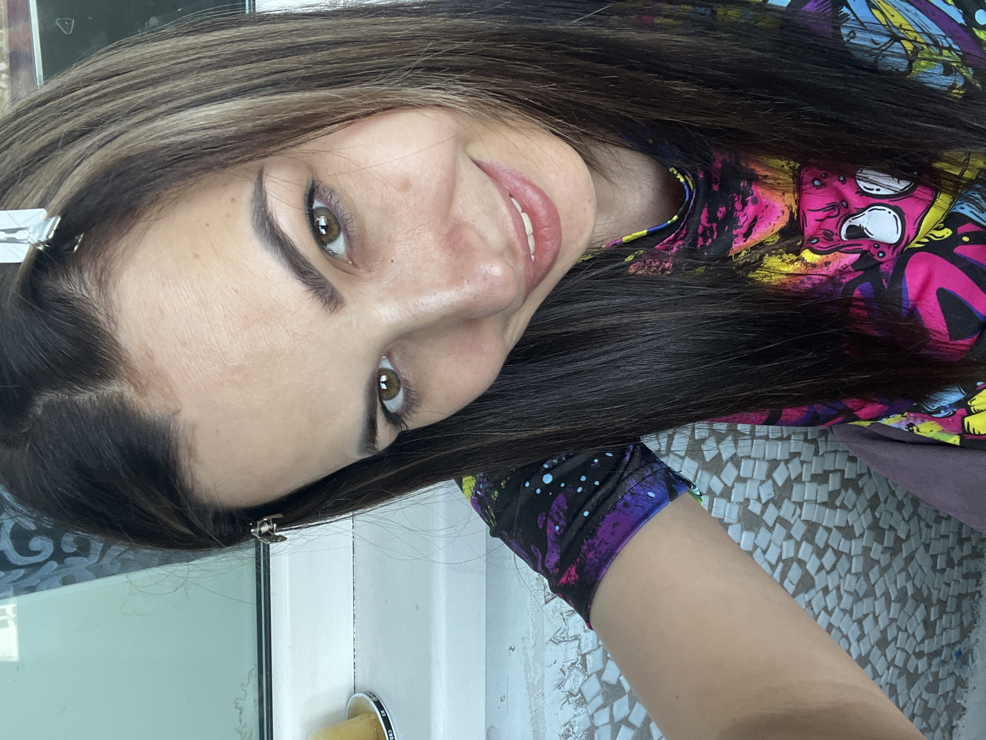 A woman with long, dark hair and light eyes is smiling at the camera. She is wearing a colorful shirt with cartoon-like images. There are two hair clips in her hair near the part. The background includes a tiled wall and a glass surface. The location is unknown.