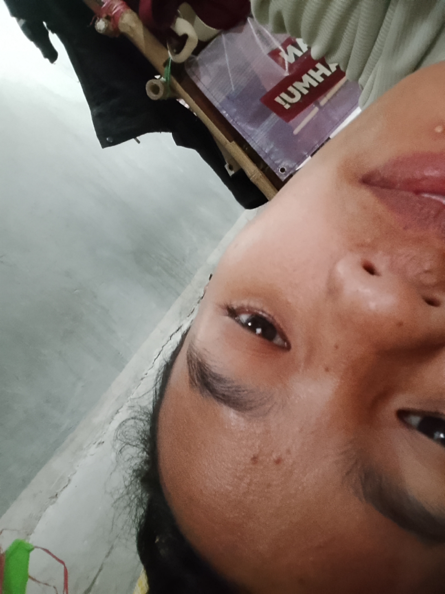 The image is a close-up, upside-down selfie of a person's face, with their eyes and mouth visible. The person's skin appears to have some blemishes and is slightly flushed. In the background, out of focus, there is a grey wall and some objects that appear to be part of a makeshift structure, possibly bamboo poles bound with red tape. A purple tarp with white lettering, possibly a logo or brand name, is also visible. The image is oriented such that the top of the person's head is towards the bottom of the frame. The exact location in Besuki, Indonesia, is not determinable from the visual information presented.