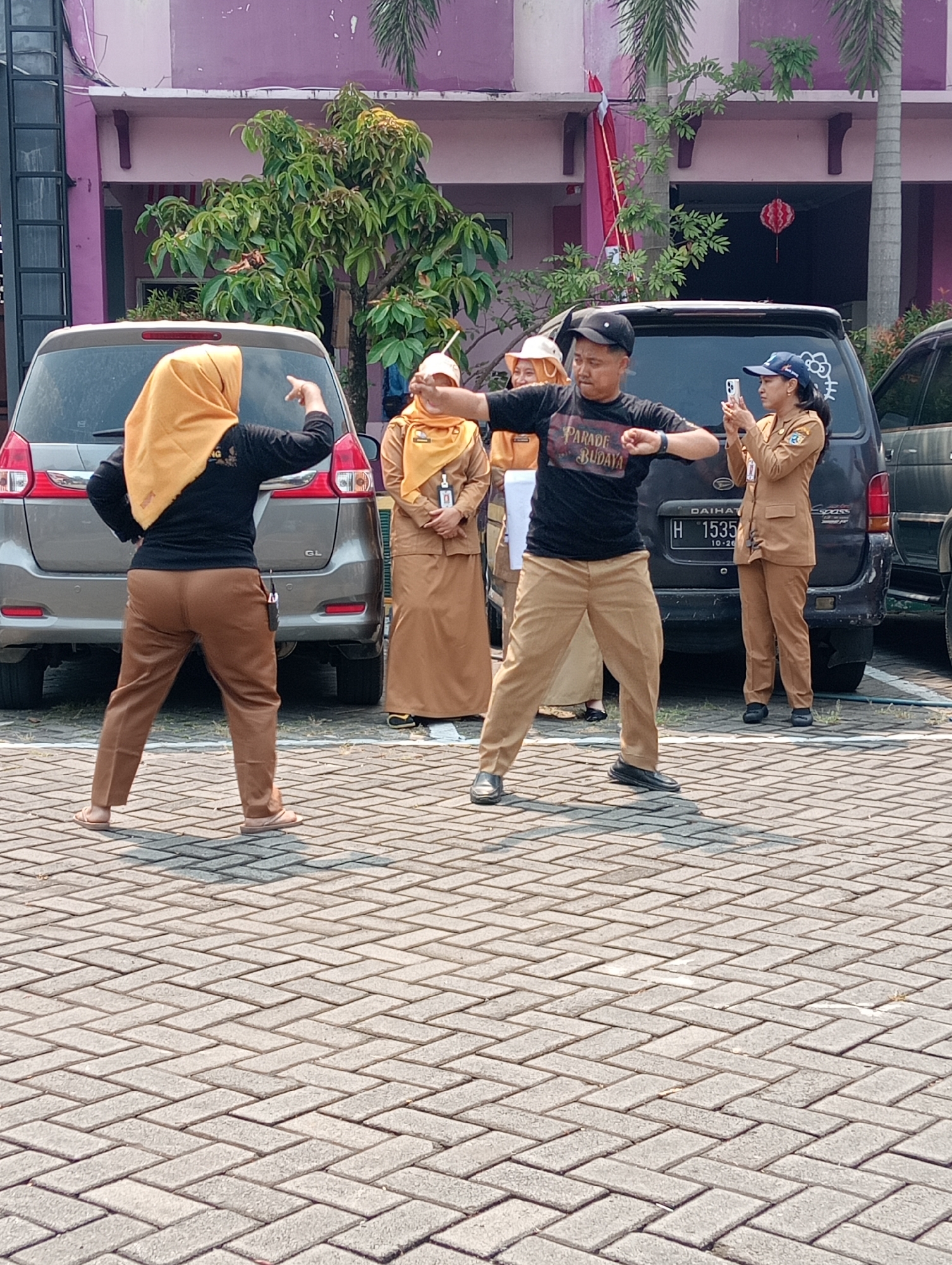 The image depicts an outdoor scene on a paved area, possibly a courtyard or parking lot, in front of a building with a purple facade. A group of individuals, predominantly women in brown uniforms, are present. Two women in the foreground are interacting: one, wearing a black shirt with "PARADE BUDAYA" written on it and a black cap, is facing right with arms outstretched, seemingly leading an activity. The other, wearing a yellow hijab and brown pants, is facing away from the camera, with her right arm raised and pointing. A third woman, also in a brown uniform and a blue cap, stands to the right, holding a smartphone and appearing to take a picture. Another woman in a similar brown uniform is visible further back, to the left of the man. Two cars, a silver SUV on the left and a dark van with a "DAIHATSU" logo on the right, are parked behind the individuals. The environment includes some greenery, palm trees, and a red lantern hanging from the building. The overall atmosphere suggests a public gathering or event. The context suggests the location is in the city of Demak, Indonesia, given the presence of individuals in uniforms commonly worn by civil servants or event staff in Indonesia.