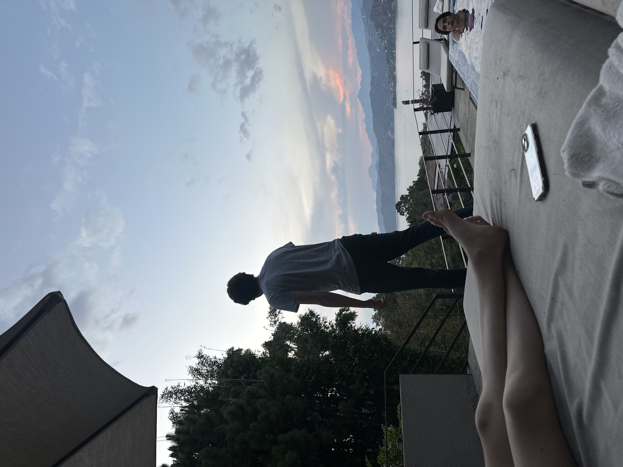 The image shows a person standing on what appears to be a balcony with a view of a lake and mountains at sunset. A woman is in a pool. The foreground contains a lounge chair with bare legs and a cell phone resting on it. An umbrella canopy is visible in the lower left. The location appears to be near a body of water with surrounding hills and trees, but the city and state are unknown.