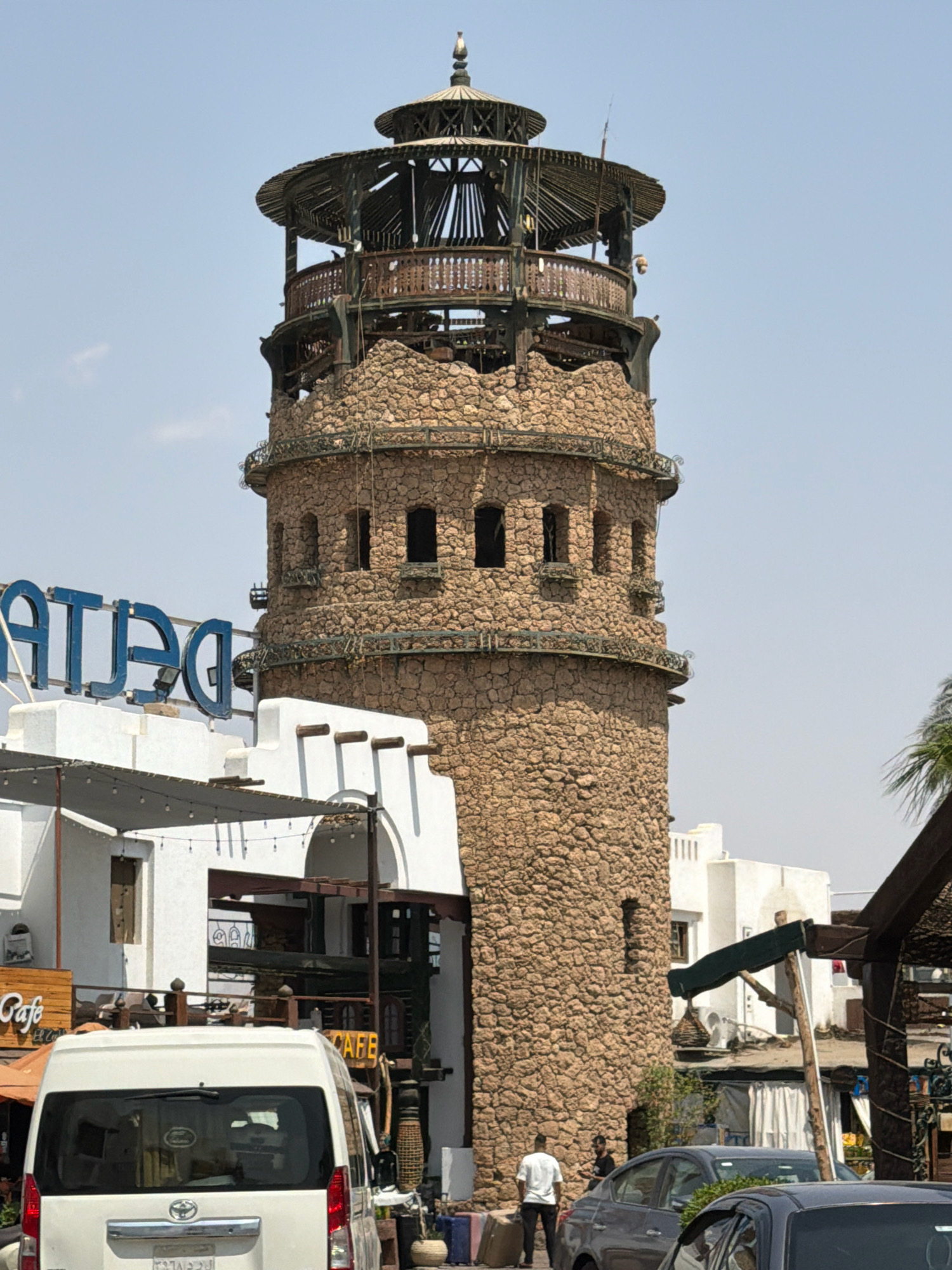 The image shows a tall, stone tower with multiple levels, topped by a wooden, open-air structure with a conical roof. The tower is constructed from rough, light-brown stones and features small, rectangular windows and decorative metalwork balconies on some levels. In front of the tower, a white building with a blue sign reading "DELTA" is visible. Adjacent to the tower and building are more structures, including a white van, a dark gray car, and a dark blue car. A few people are visible; one in a white shirt stands near the van, and another in a dark shirt stands near the cars. A palm tree is partially visible on the right side of the image. The background is a clear, light blue sky. The scene appears to be an outdoor area in Sharm El Sheikh, Egypt, likely a commercial or tourist district, given the presence of cafes and shops suggested by signs like "Cafe" and the architectural style.