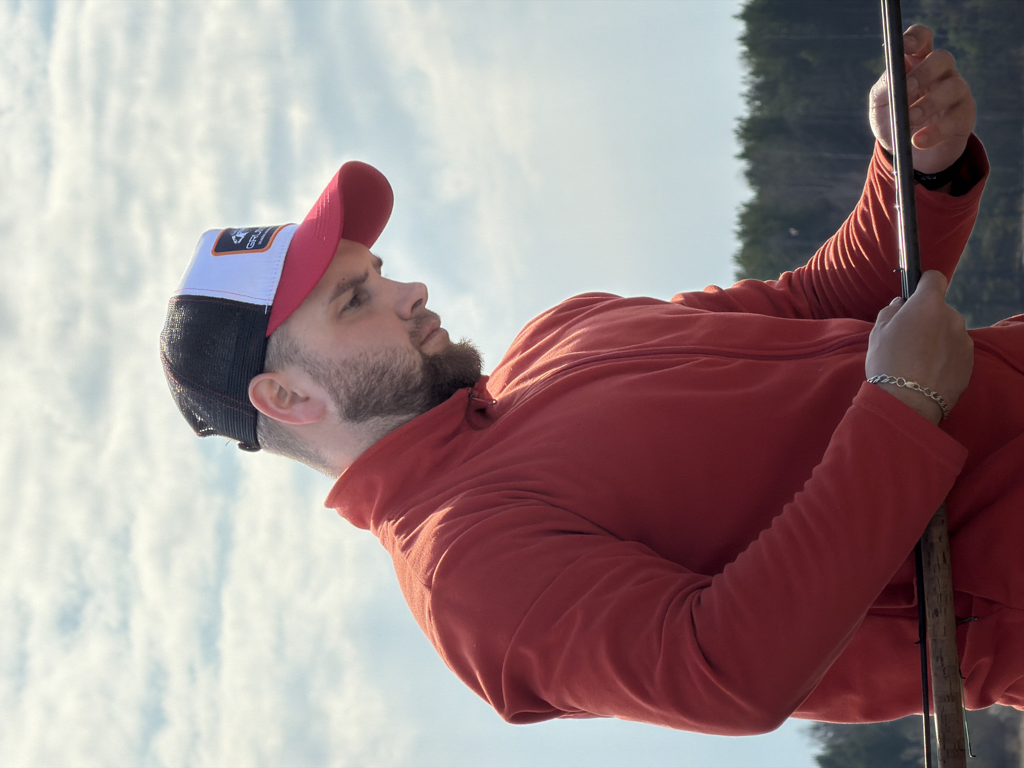 A man with a beard is holding a fishing rod. He is wearing a red jacket and a baseball cap that is red, white, and black. In the background is a lake and a forest. The sky is visible above with some clouds. The photograph may have been taken in Chaykovsky, Russia.