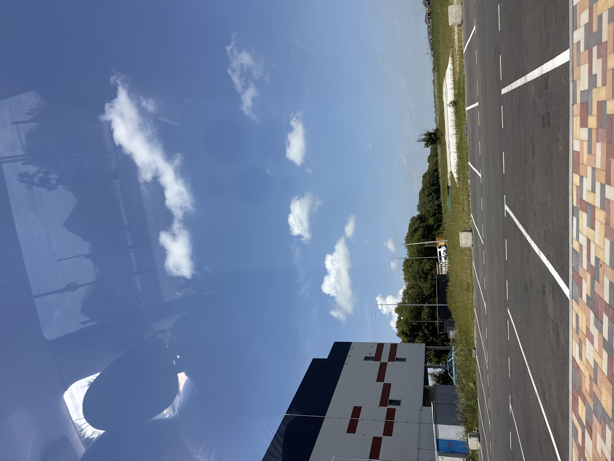 The image depicts an outdoor scene, likely a parking lot, with a large industrial building on the left. The building is white with dark blue and red horizontal accents. In the foreground, there is a paved parking lot with white lines marking parking spaces. Beyond the parking lot, there is a grassy area with scattered trees and some low-lying structures or debris in the distance. The sky is blue with scattered white clouds, and reflections of trees and possibly a vehicle are visible on a glass surface in the upper left portion of the image, suggesting the photo was taken from inside a vehicle or through a window. The location is specified as Mikhaylovsk, Russia.