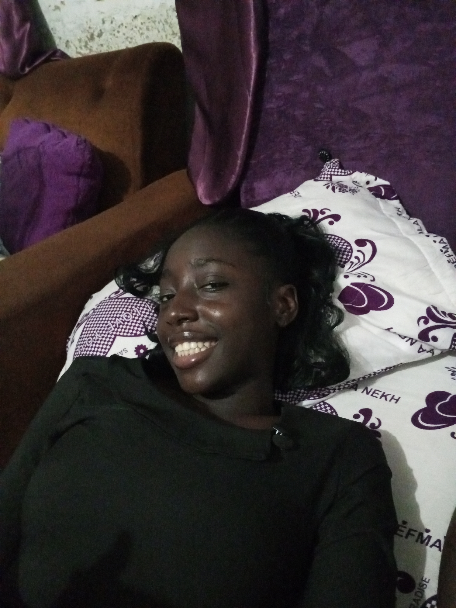 A young woman with dark skin and short, dark hair lies on a white pillow with purple patterns. She is wearing a dark-colored long-sleeved shirt and smiling. The background includes a purple and brown couch. The location is unknown and cannot be determined from the image.