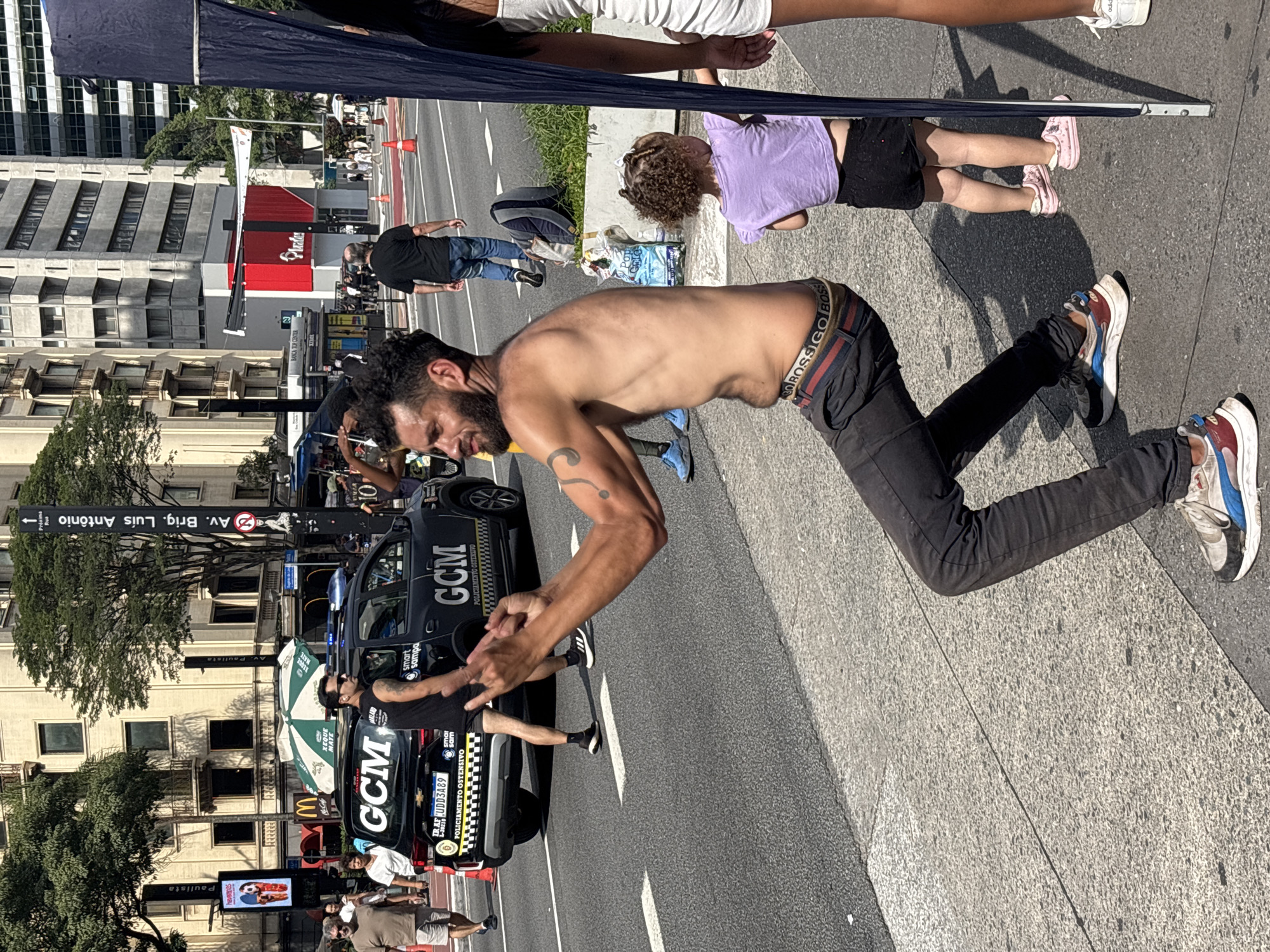 The image, rotated 90 degrees counter-clockwise, depicts a busy street scene in São Paulo, Brazil. In the foreground, a shirtless man wearing dark jeans and white-blue-red sneakers bends at the waist, his hands clasped together. A child in a purple shirt and black shorts lies on the sidewalk nearby. In the background, there are people, a black police car marked "GCM" (Guarda Civil Metropolitana), and street signs including "Av. Brig. Luís Antônio" and "Paulista". A McDonald's sign and advertisements are visible in the background. The scene appears to be captured during daylight hours.
