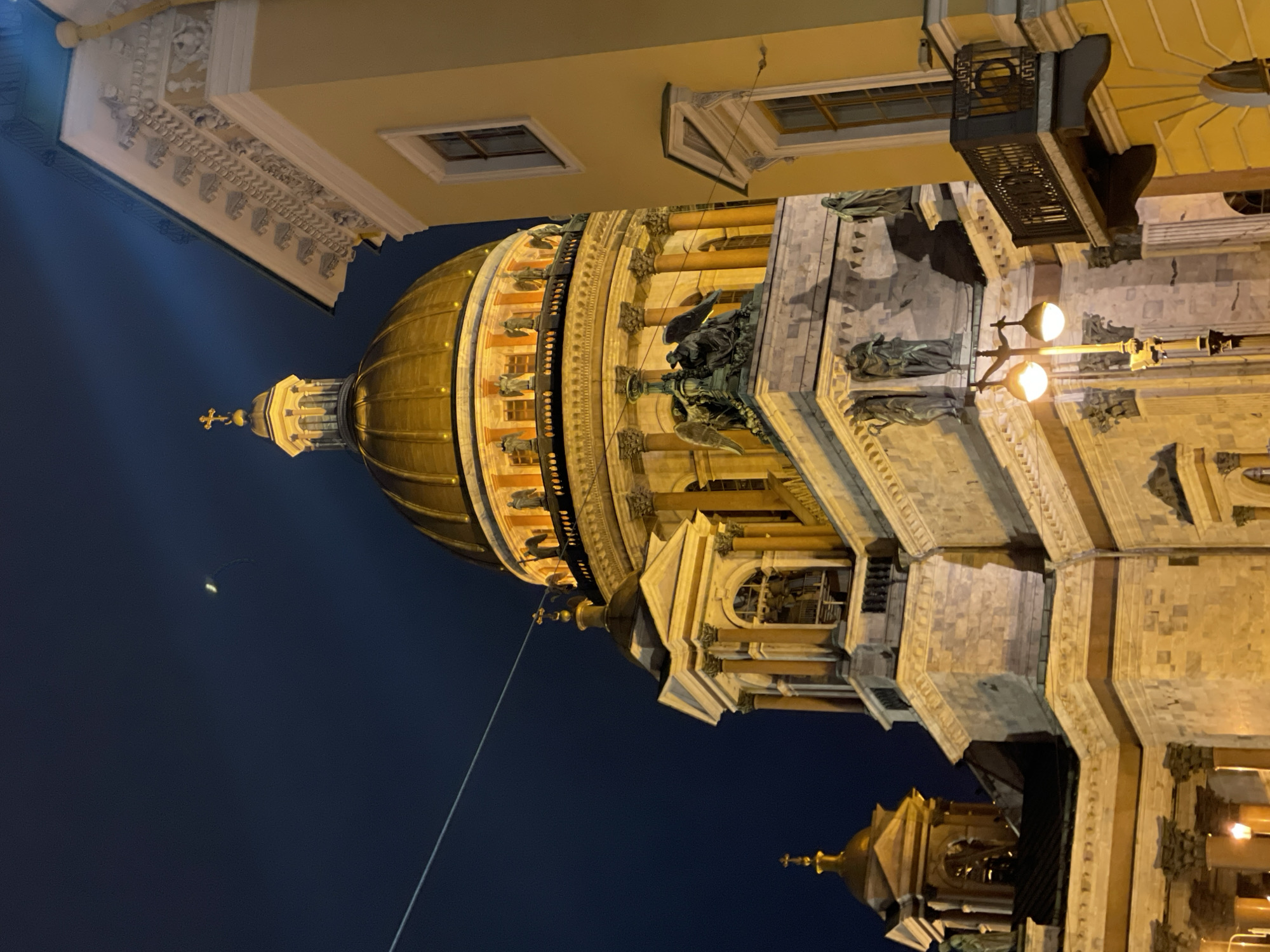 The image shows a low-angle view of the Saint Isaac's Cathedral in Saint Petersburg, Russia, at night. The main dome is illuminated, and the surrounding structures of the cathedral are visible, showcasing architectural details such as columns, statues, and ornate carvings. The sky is dark, suggesting it is nighttime. A building is adjacent to the Cathedral and the photo is taken from below.